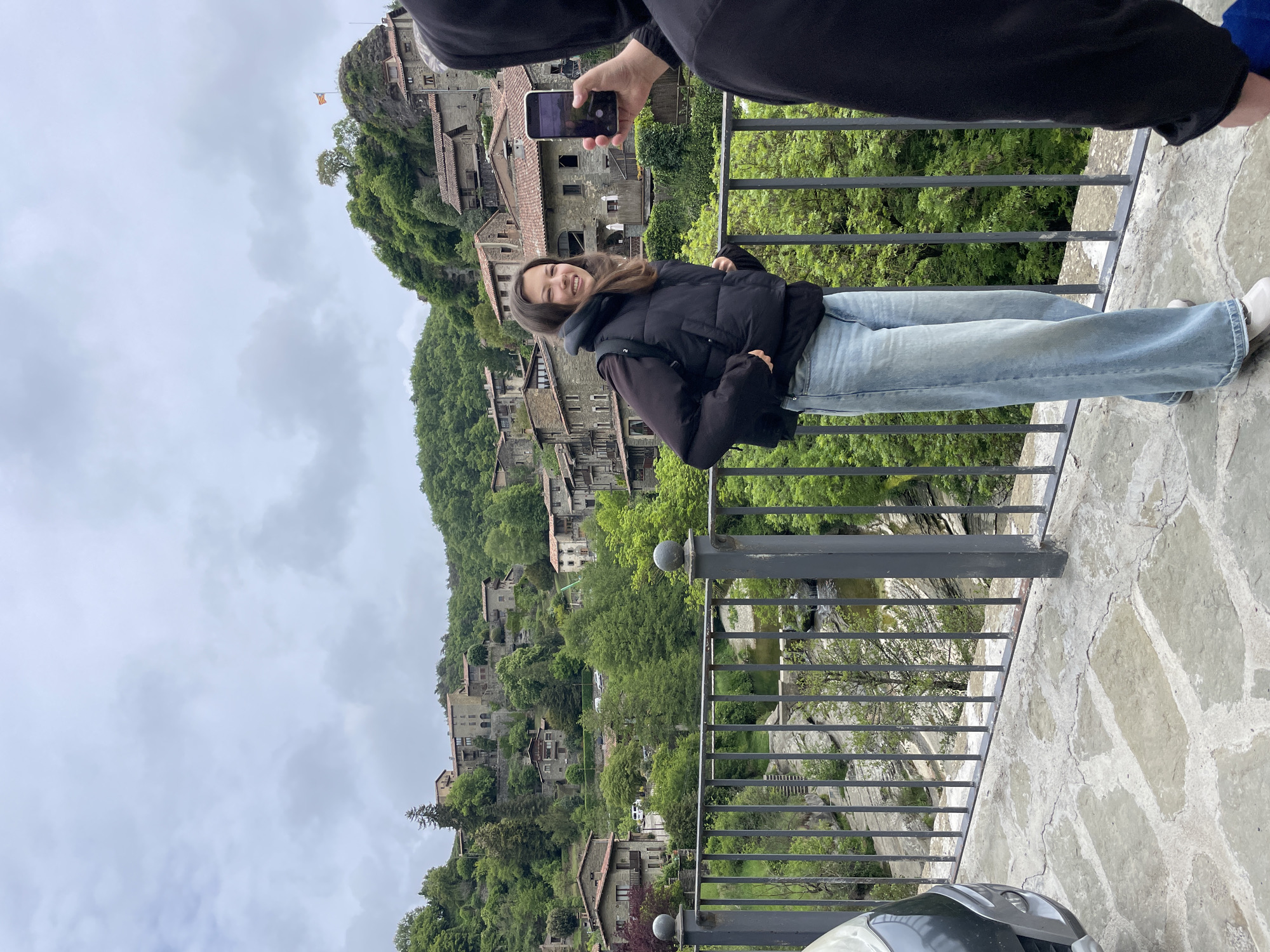 A vertical outdoor shot depicts a young woman posing on a stone bridge. A second person, partially visible, is holding a cell phone to take her picture. The woman is wearing a black puffer jacket and blue jeans. She smiles at the camera. The bridge has a gray metal railing. Below the bridge is a river or stream. Behind the woman, the hillside is covered with stone buildings and green trees. The sky is overcast. This is a view of Rupit i Pruit, Spain.