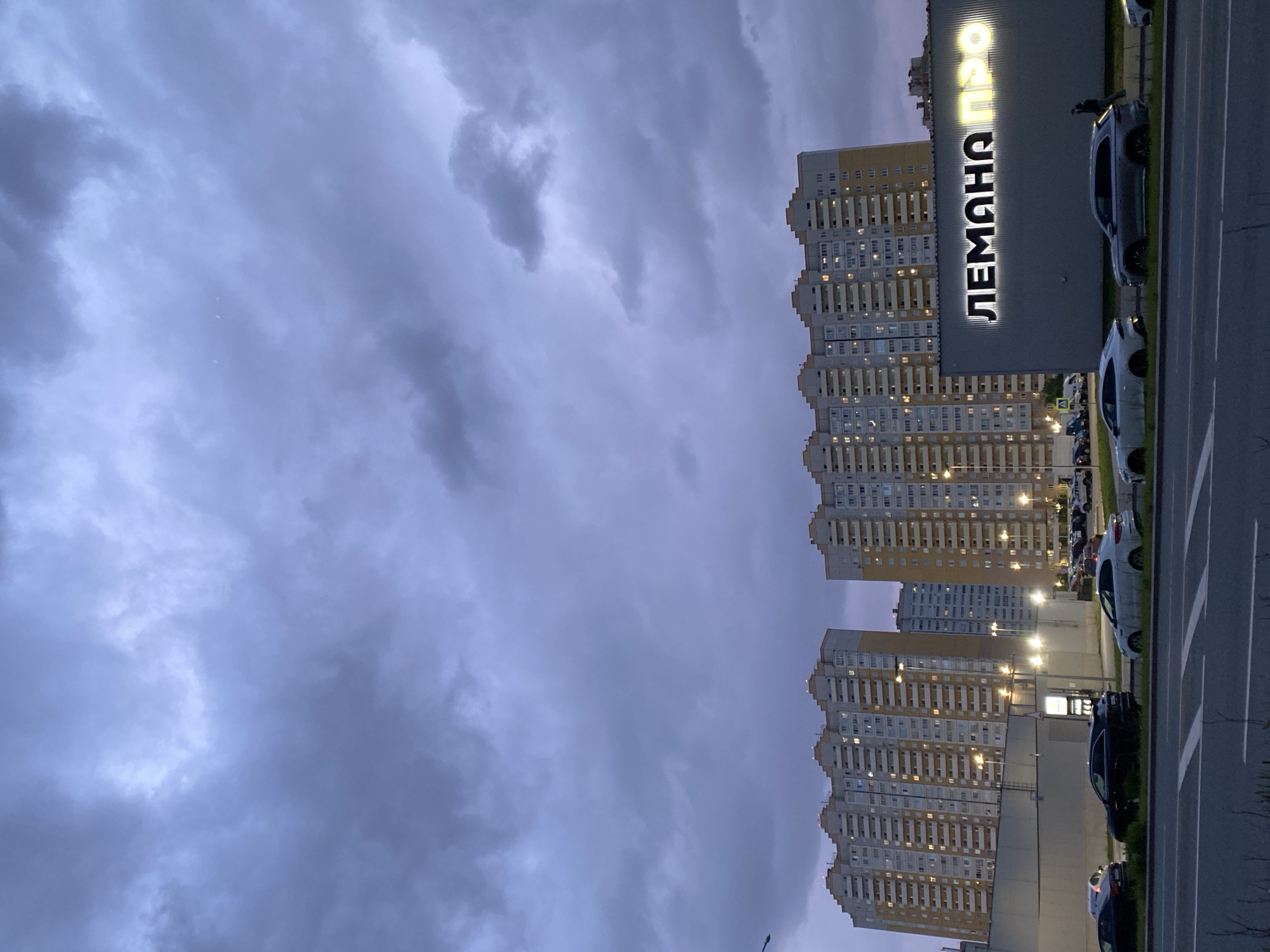 The image captures a twilight scene in Saint Petersburg, Russia, featuring the LEMAN PRO hypermarket, and residential buildings. The sky is dominated by heavy, overcast clouds. To the right are several high-rise apartment buildings with lit windows. Adjacent to the buildings is the LEMAN PRO hypermarket with its name illuminated in Cyrillic script. A parking lot filled with vehicles is visible in front of the hypermarket. A road with lane markings is in the foreground, next to the parked cars.