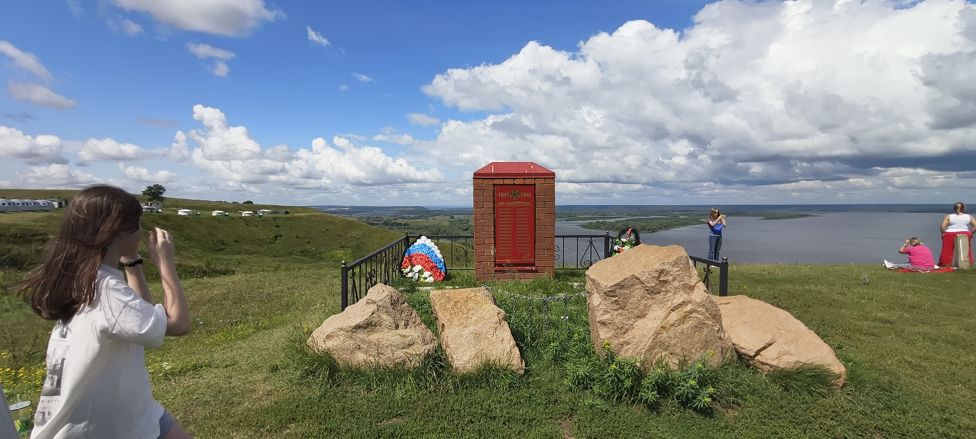An eye-level, medium outdoor shot shows a memorial monument on a grassy hill overlooking a wide river on a cloudy day. A red brick monument with a red roof is in the center, displaying a list of names on a red plaque. A wreath of red, white, and blue flowers sits in front of the monument. Three large rocks are in the foreground.
To the left of the monument, a young woman with long brown hair is taking a photo with her phone. She is wearing a white t-shirt and gray shorts.
In the background, across the river, the landscape is green and rolling. Several white yurts or tents are visible on a distant hill. On the right side of the frame, two women are sitting on the grass, one in a pink shirt and the other in a white shirt and red skirt. Another woman stands near the railing, looking out at the river. The sky is partly cloudy with scattered white clouds.