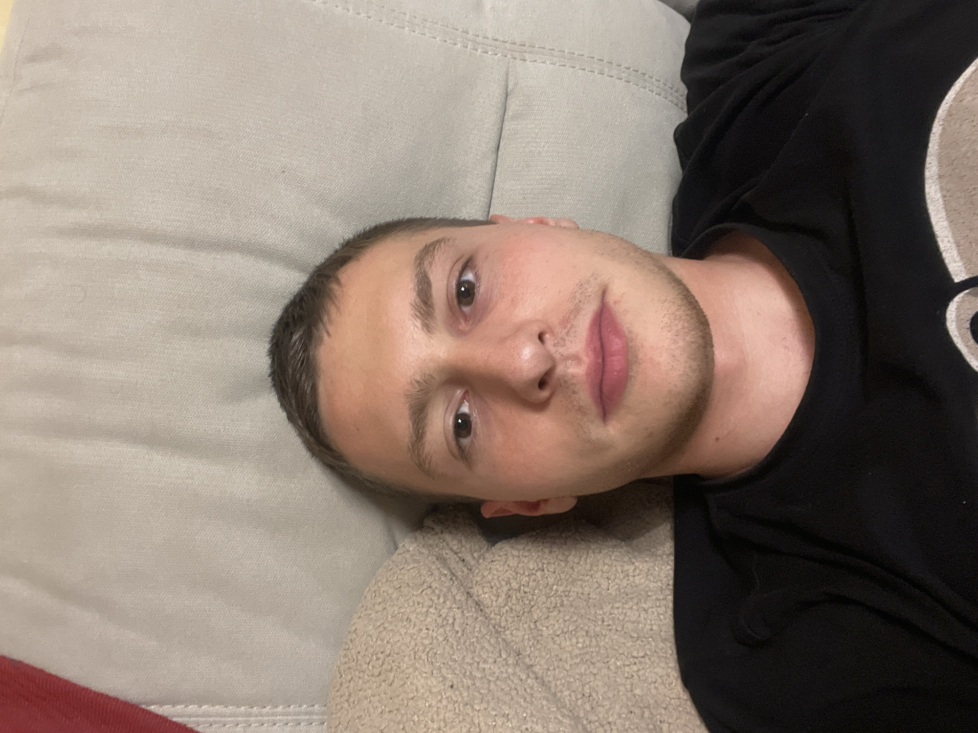 A young, light-skinned male lies on a beige colored sofa. He wears a black t-shirt. The location is unknown; no evidence of the city of Burgas, Bulgaria is visible.