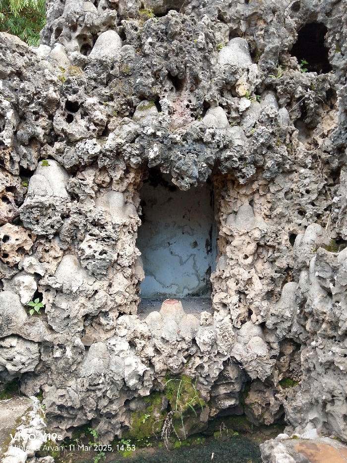 The image shows a close-up of a rocky wall with a small opening, located in Cirebon, Indonesia, at the coordinates -6.736 latitude and 108.544 longitude. The wall is composed of grey and beige, porous rock, possibly limestone, with various indentations and formations. Some green plants are visible at the bottom of the image, along with a small pool of water. The opening in the wall reveals a white concrete or plastered surface with a small ledge on which a red object rests. The image is taken at a close distance to the wall and appears to be a still shot taken during the day. The image is well-lit, but not overly bright. There is a small watermark in the bottom left corner of the image.