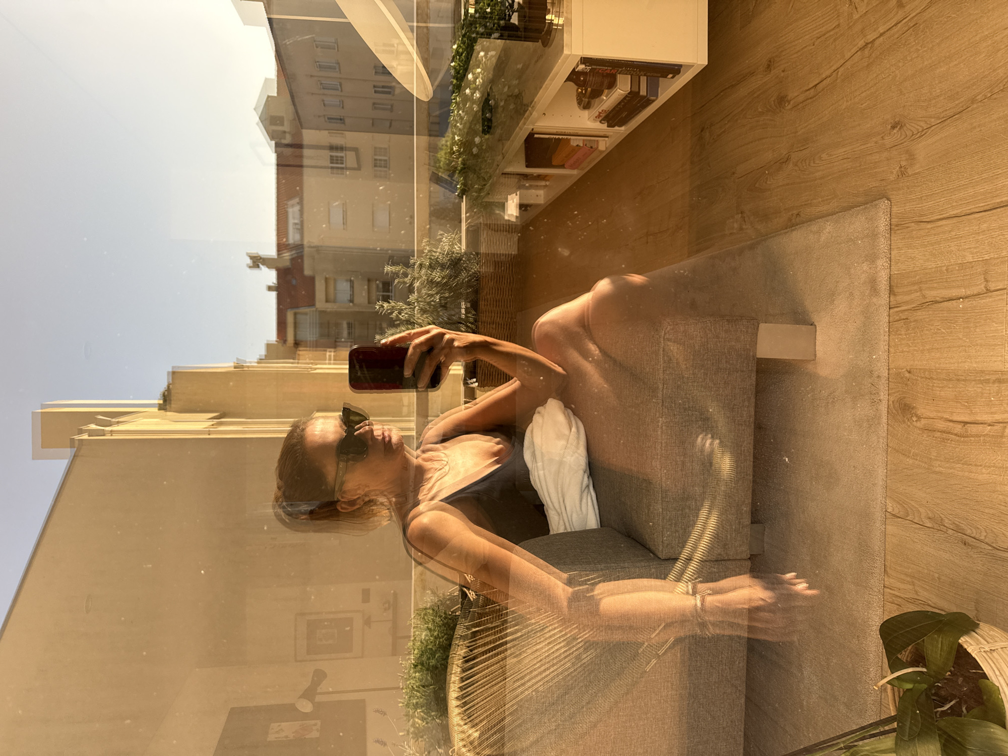 The media file is a reflection of a woman taking a selfie in a window. The woman is sitting on a chair, wearing sunglasses and a black swimsuit. She is holding a phone in her hand, and her body is partially visible. Behind her, the reflection shows an interior scene with a wooden floor, a rug, and a shelf with books and plants. The reflection also shows an exterior scene with buildings and a clear sky, suggesting it is daytime. The overall emotion conveyed is one of relaxation and self-confidence. The image is likely taken in Vila Nova de Gaia, Portugal, as indicated by the location context.