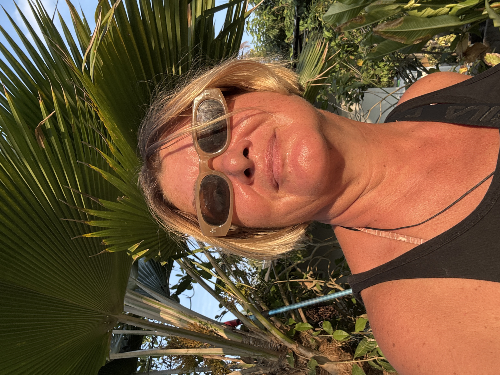A woman with blonde hair and sunglasses is taking a selfie in Baan Mae Nam, Thailand. She is wearing a black top. A large fan palm is visible in the background. Other greenery, including trees and shrubs, is also present. A blue pipe is visible behind the woman. The sky is partly visible between the leaves.