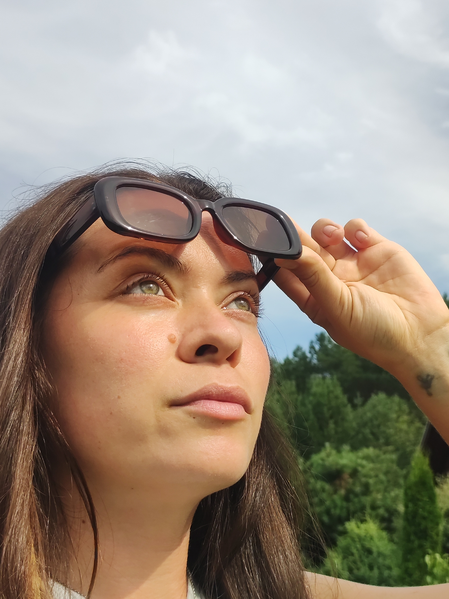 A young woman with medium-length brown hair and green eyes is looking up and to the right, her sunglasses perched on her forehead. She is wearing a light-colored shirt. Her right hand is raised, holding the sunglasses. The background is a cloudy sky and a line of green trees, suggesting an outdoor, possibly rural or park setting. There is no indication of the city of Фоки, Russia, in the image.