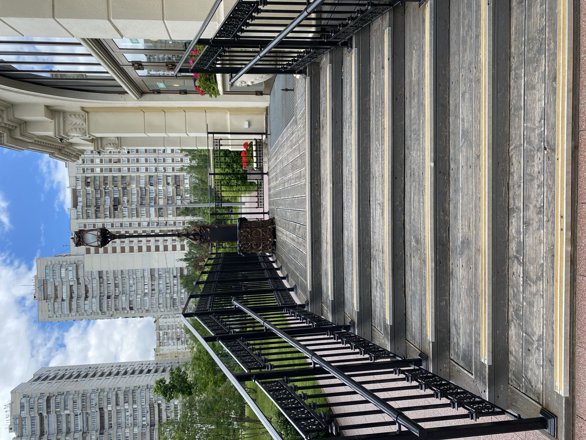 The image shows a set of wooden steps with black metal railings leading up to a building. The steps appear to be outdoors, with tall apartment buildings and greenery visible in the background. The location is identified as Khimki, Russia, based on the presence of specific architectural details and urban layout characteristic of the city. There are no people visible in the image. The steps are made of planks of wood, with lighter-colored strips running parallel to the edges of the planks. The metal railings are ornately designed and appear to be constructed from black iron. The architectural style of the buildings in the background is typical of Soviet-era housing blocks.