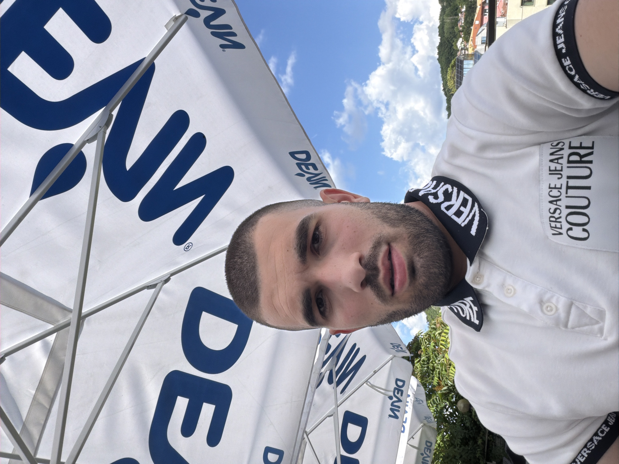 A man with short dark hair and a beard is taking a selfie outdoors. He is wearing a white polo shirt with black and white trim on the collar and sleeves that reads "VERSACE JEANS COUTURE". He is positioned in the foreground, with a large white umbrella with blue lettering behind him. The umbrella has the word "DENIM" written in large blue letters. The background shows a bright blue sky with scattered white clouds, and a town with buildings nestled on a hillside with green trees. The man's expression is neutral. The lighting suggests it is daytime.