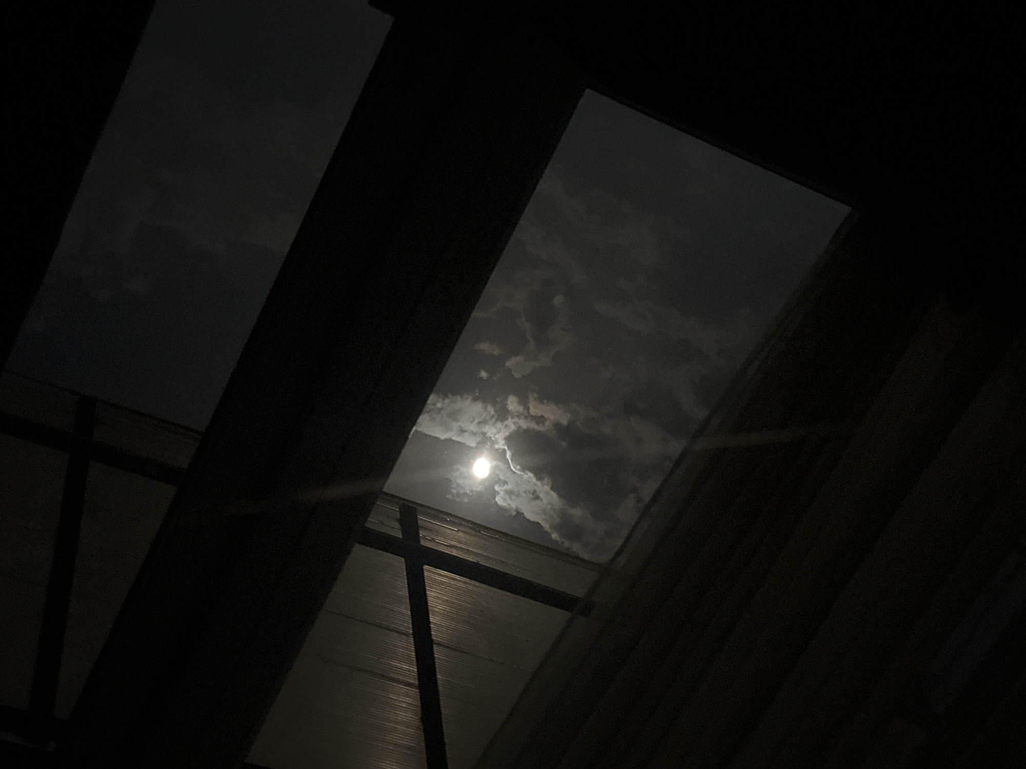 The image depicts a nighttime view of the sky, specifically a cloudy night with the moon visible through an opening in a structure, possibly a skylight or window. The frame consists of dark structural elements that appear to be part of a building's interior. The moon is a bright orb surrounded by dark clouds. The city of Yerevan, Armenia, is not verifiable from this image. No people, subjects, objects, or actions can be described further without additional details.