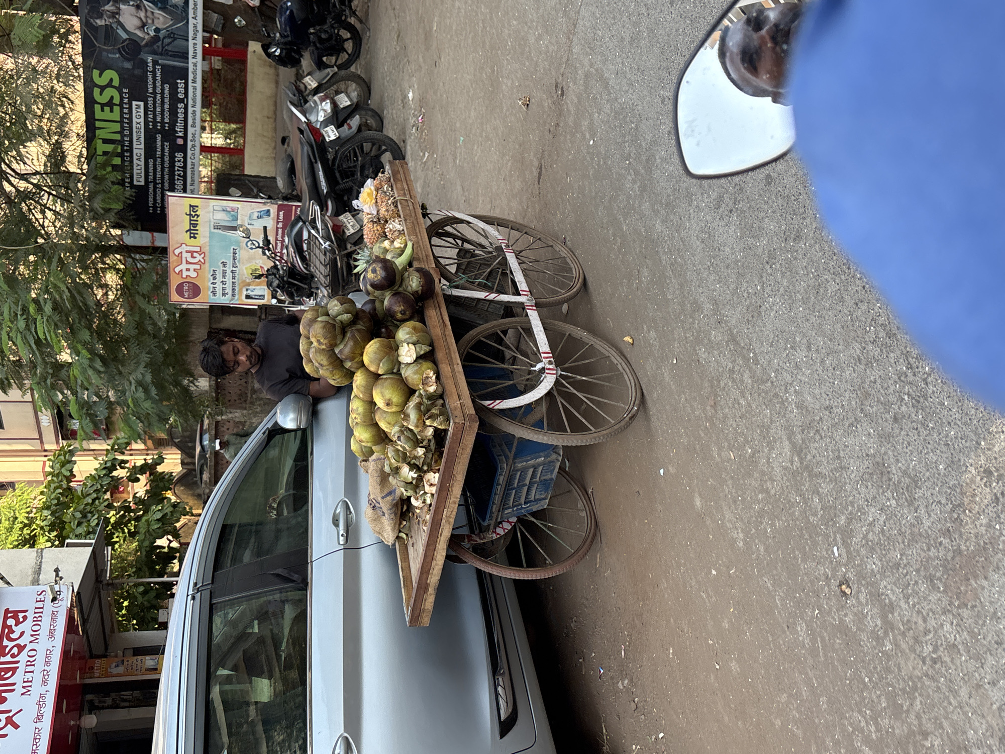 A street scene in Ambernath, India, shows a vendor selling coconuts and pineapples from a three-wheeled cart. The cart is positioned between a parked silver car and the curb. The vendor, a man in a dark shirt, stands near the cart. In the background, there are motorcycles parked near a building with signs for "Fitness" and "Metro Mobiles" at Int. Namaskar Co.Op.Soc., Beside National Medical, Navre Nagar. A side mirror is visible in the foreground.