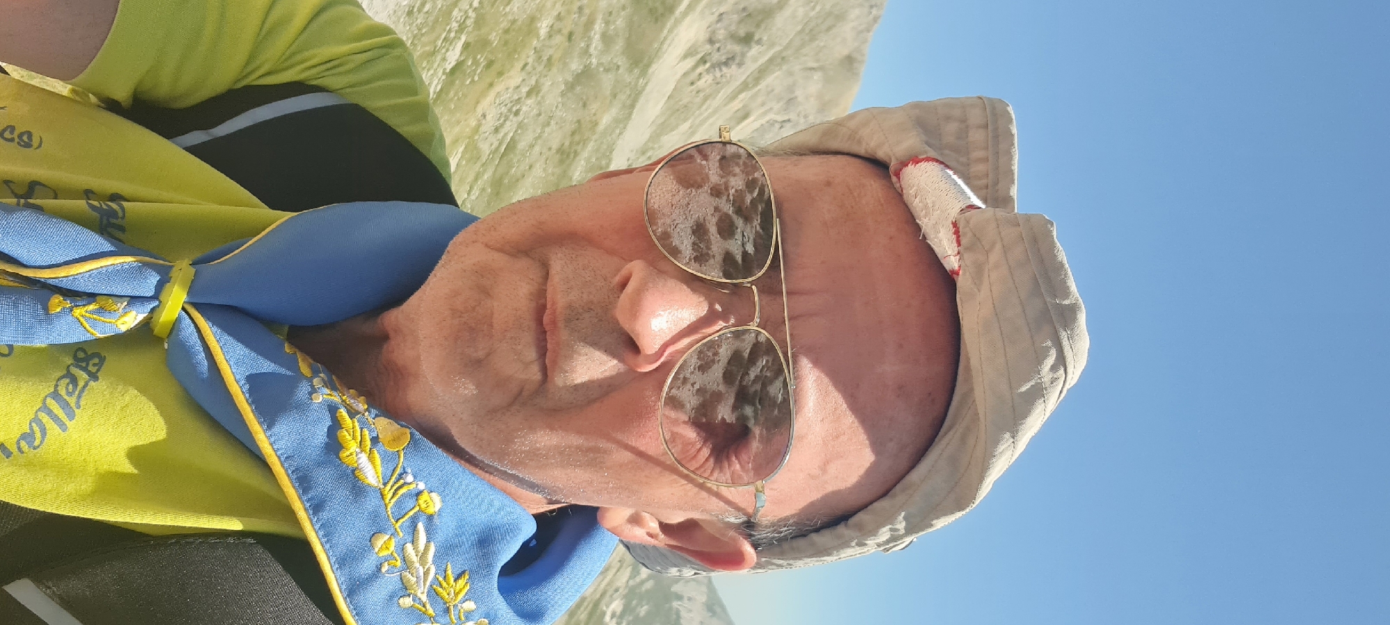 This is a selfie taken outdoors with a clear blue sky above. The subject is a man wearing a light-colored cap, aviator-style sunglasses, a bright yellow t-shirt, and a blue neckerchief with yellow embroidery. The background shows a mountainous landscape, typical of the Gran Sasso mountain range near L'Aquila, Italy, characterized by rocky slopes and sparse vegetation. The man appears to be in good health and is looking directly at the camera. The overall impression is of a person enjoying an outdoor activity in a scenic, elevated location.