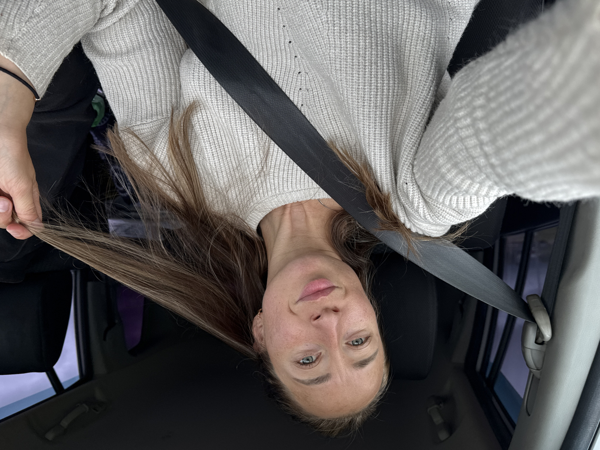 The image features a single female individual seated in the passenger side of a vehicle, appearing to take a selfie. She is positioned facing forward and slightly to her right, looking directly at the camera. Her hair is long, light brown, and styled loosely, with some strands pulled forward over her left shoulder, and a section held by her right hand near her chin. She has light-colored eyes, a light complexion, and visible freckles across her nose and cheeks. Her lips are closed in a subtle smile. A small, gold-colored hoop earring is visible on her right earlobe.
She is wearing a light, possibly off-white or light gray, ribbed knit sweater. A black seatbelt strap crosses diagonally over her chest and left shoulder, securing into a mechanism on her left side. The seatbelt's height adjuster is visible on the vehicle's A-pillar to her left. On her left wrist, a thin black string bracelet with a small metallic bead is visible.
The interior of the vehicle appears dark, with black upholstery visible on the seats behind her and to her right. The overhead console and a grab handle are partially visible in the upper right background. Through the side windows, an overcast or low-light exterior is suggested by muted colors and soft light, with no distinct landscape features. A faint purple object is discernible between the front and rear seats on the far right side of the frame.
The image provides no external visual cues or contextual information to identify the specific location as Karagay, Russia.