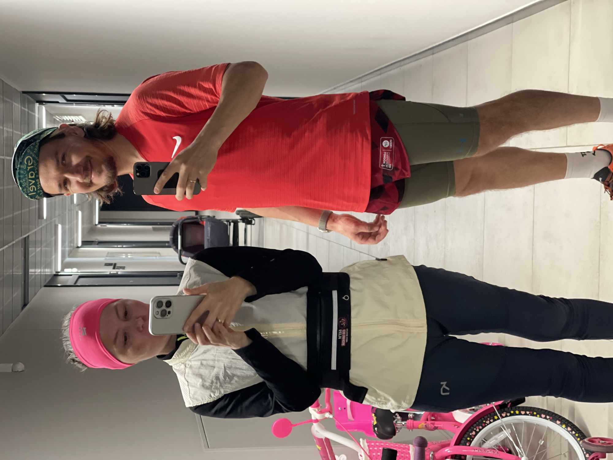 Two people are posing for a selfie in a hallway. The person on the right is a man wearing a green hat, a red Nike t-shirt, green shorts, and orange running shoes. The person on the left is a woman wearing a pink headband, a white vest, a black long-sleeved shirt, and black leggings. A pink bicycle is visible in the foreground on the left. The hallway has white walls and a row of fluorescent lights on the ceiling. There are chairs and closed doors visible in the background. The location is unspecified and not discernable from the image.