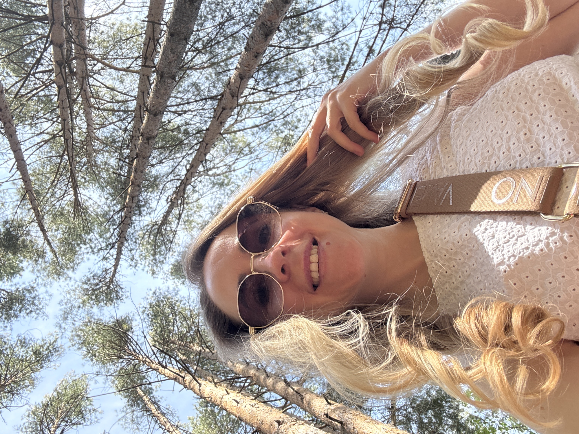 A woman with blonde hair, wearing sunglasses and a white blouse, is posing in an outdoor setting. The photo is taken from a low angle, looking up at the woman, with trees and a clear blue sky visible in the background. The shoulder strap is visible on the white blouse. Given the location, Chaikovsky, Russia, the environment appears to be a forest.