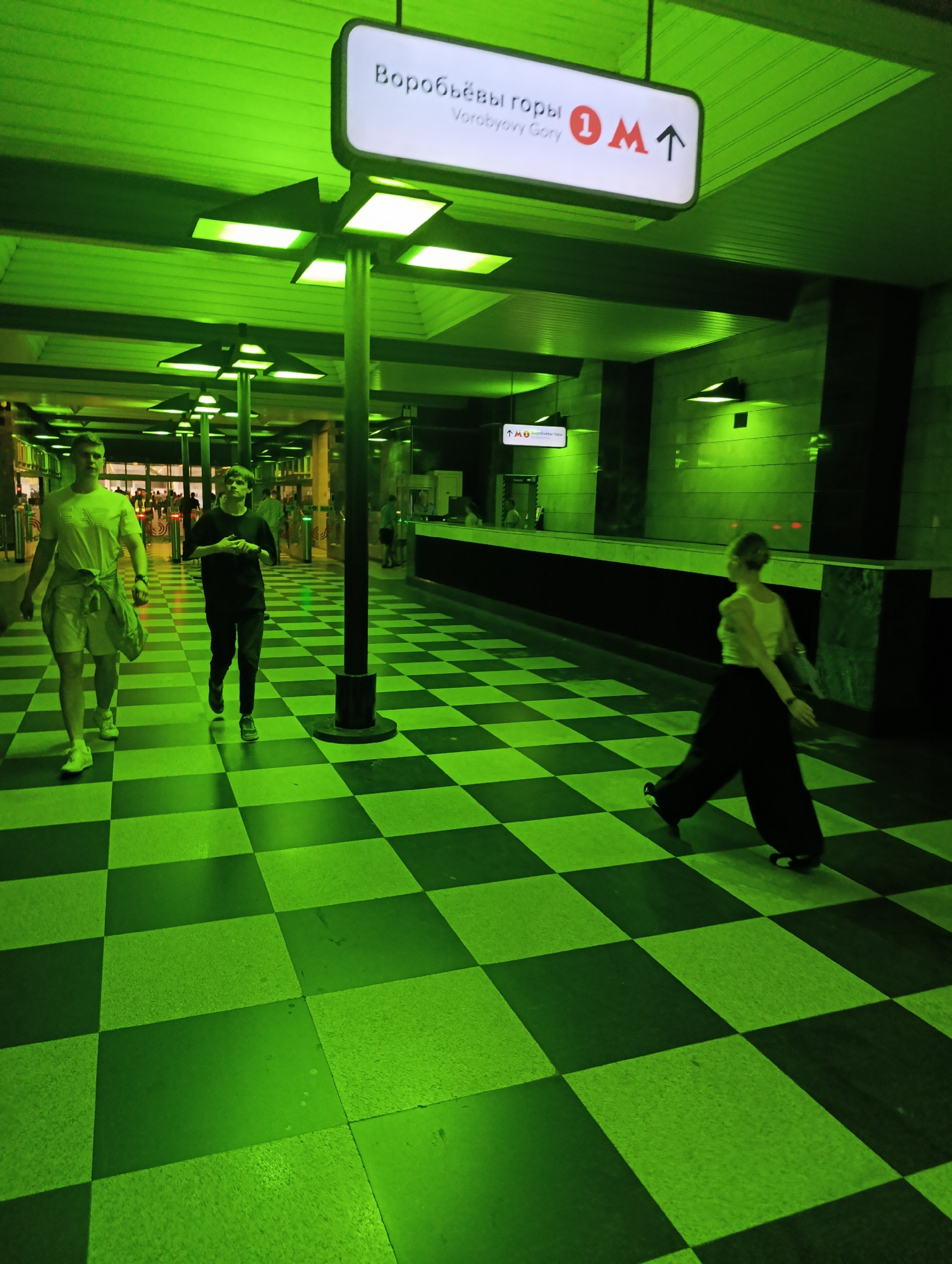 The image displays the concourse of the Vorobyovy Gory metro station in Moscow, Russia, characterized by a pervasive, monochromatic green illumination emanating from square ceiling lights. The floor features a distinctive large-scale checkerboard pattern of alternating dark green and light green squares.
A prominent overhead white sign, suspended from a dark central pillar, displays "Воробьёвы горы" in Cyrillic, "Vorobyovy Gory" in English, and a red "1 M" logo, indicating Metro Line 1, accompanied by an upward arrow. A smaller, similar sign is visible further down the concourse on the right wall.
Three individuals are in motion within the space. On the left, a man wearing a light-colored t-shirt, light shorts, and a jacket tied around his waist walks towards the right side of the frame. Trailing him, a younger person dressed in a dark t-shirt and dark pants also walks right. On the right, a third person, wearing a light-colored top and dark, wide-leg pants, walks away from the viewer towards the left.
The concourse is supported by dark pillars featuring square light fixtures. In the background, beyond the main subjects, several turnstiles are visible, with additional blurred figures of people near what appears to be an entrance or exit. A long, dark counter or ledge extends along the right wall, which, like the ceiling, exhibits a textured or ridged surface.