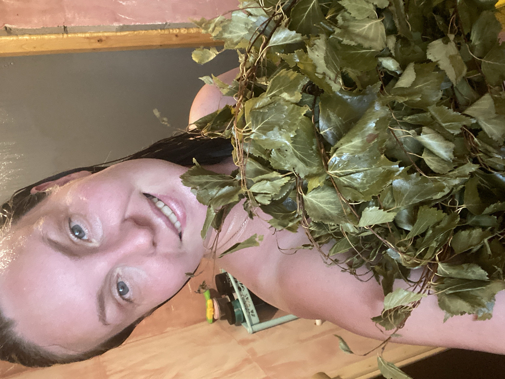 The image shows a woman holding a bunch of birch branches. She is in what appears to be a sauna or steam room, indicated by the wooden walls and the steam visible on her skin. Her hair is wet, and she has a smiling expression. In the background, there is a shelf with various containers, possibly toiletries. The overall lighting suggests an indoor environment, likely humid and warm, typical of a sauna. The woman's attire is not clearly visible, but she is partially covered by the birch branches.