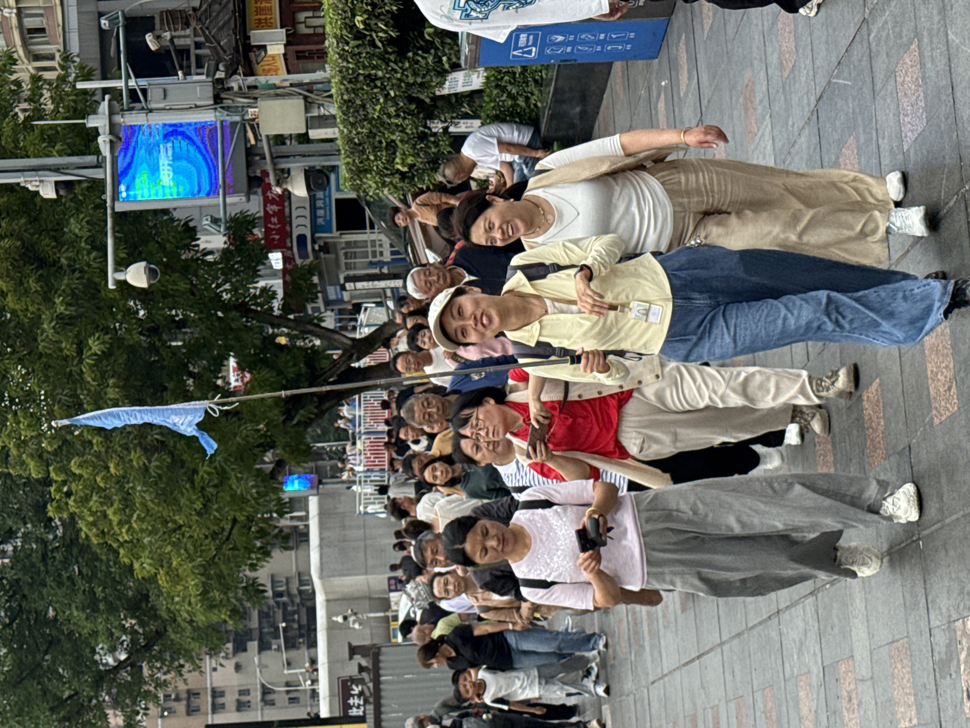 A group of people are walking on a paved sidewalk in the city of Yuzhong District, China. They are walking in a straight line. Some of them are smiling. One is holding a blue flag on a pole. There is a tall building in the background. There are trees to the left of the crowd and buildings to the right. There is also a digital display on a pole. Several signs and billboards are nearby.