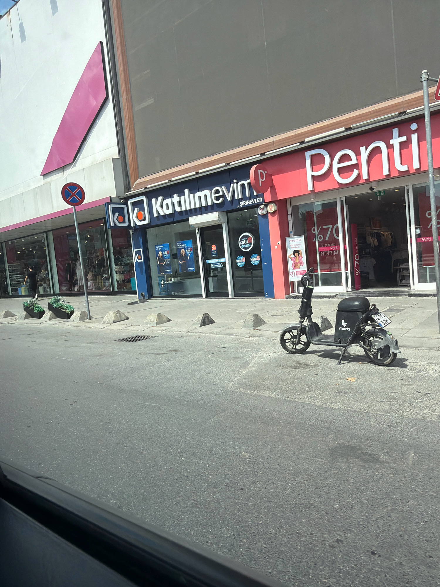 The image depicts a street scene in Bahçelievler, Turkey, featuring storefronts and a parked electric scooter. On the left, a building displays a large, abstract pink geometric shape on its facade. Next to it is a store with blue and white signage that reads "Katılımımevim" with the subtitle ".ŞİRİNEVLER" below it. The storefront windows showcase merchandise and promotional posters.
To the right of "Katılımımevim" is a store with bright red signage displaying "Penti." This store also has large glass windows revealing clothing displays and promotional signs, including one with "%70" and text in Turkish.
In front of the stores, a dark-colored electric scooter is parked on the sidewalk. The scooter has a black seat and a license plate with the characters "34 FBZ 628". The brand "martı" is visible on the scooter's rear storage compartment. The street in the foreground is asphalt, with a gutter and a manhole cover visible. Several concrete bollards are placed along the sidewalk. The environment suggests a commercial area in an urban setting.