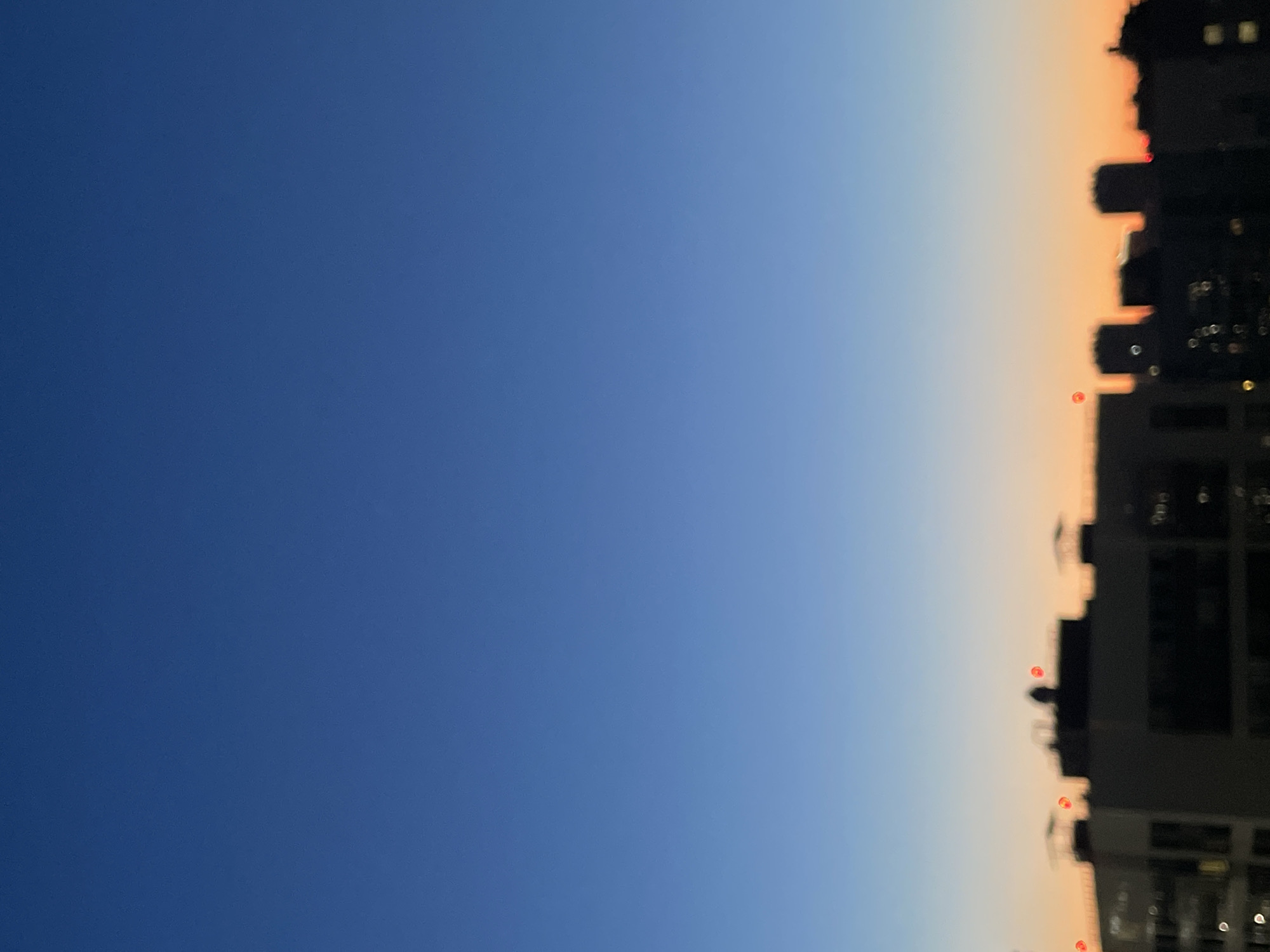 The image depicts a cityscape at dusk. The sky transitions from a deep blue at the top to a pale orange near the horizon. Silhouetted buildings of varying heights form the lower portion of the frame. Several of the buildings have illuminated red lights on their rooftops. The image is out of focus, giving the scene a soft, blurred appearance. There is no indication of the specific location within the city of Sinyavino, Russia. There are no people or discernible actions visible in the image.