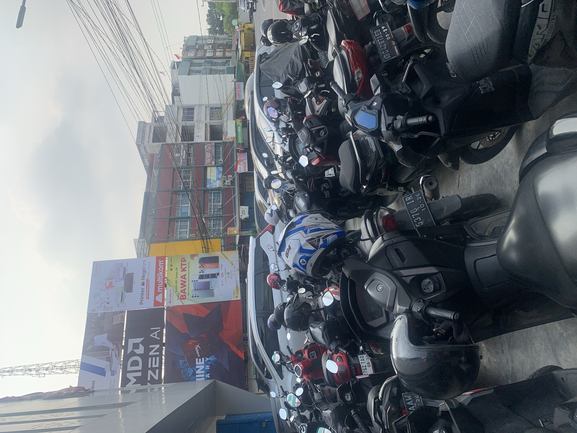 This is a low-angle, medium shot photograph taken outdoors, likely in the city of Palembang, Indonesia, given the presence of Indonesian license plates and an advertisement in Indonesian. The scene is a parking lot filled with numerous parked motorcycles and several cars. In the background, multi-story buildings with storefronts and windows are visible, some displaying advertisements. Electrical wires are strung across the upper portion of the image. The lighting suggests it is daytime, possibly overcast, with a diffused sky. The focus is on the motorcycles, many of which have helmets placed on their seats or handlebars. The overall impression is of a busy urban environment.