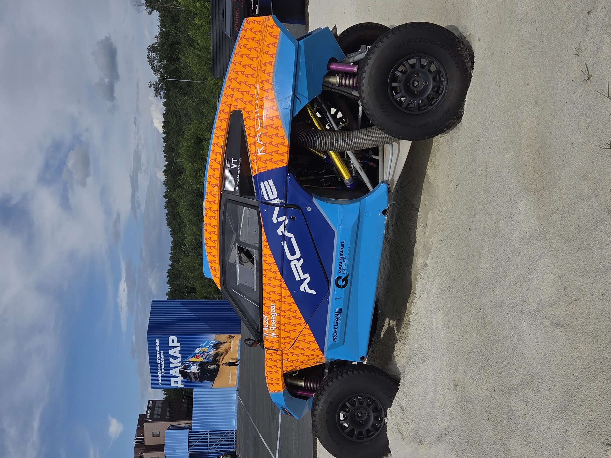 The image depicts a rally car, likely a Dakar Rally participant, on a sandy surface. The car is predominantly blue and orange, with a repeating abstract pattern of orange "A" shapes on the orange sections. The car features large, off-road tires, exposed suspension components with colorful springs, and aerodynamic bodywork. In the background, a large blue structure with "DAKAR" prominently displayed in white letters is visible, alongside buildings and a dense forest. This suggests the location is likely an event or exhibition related to the Dakar Rally. While the city of Yekaterinburg is mentioned, the image itself does not contain any explicit markers to confirm this specific location. There are no people clearly visible in the immediate foreground or interacting with the car. The overall environment appears to be an outdoor setting with a cloudy sky.