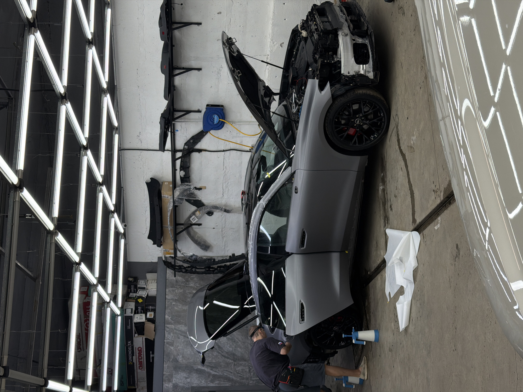 A gray car undergoing maintenance is positioned in a garage-like setting under bright lighting, and a person is working on the vehicle. The car has its hood open and front panels removed, revealing the engine components. The individual is near the front of the car and is dressed in a dark-colored shirt and shorts. The garage has a concrete floor, and car parts are stored on shelves against the wall. A blue hose reel is mounted on the wall above the vehicle. The reflection of the lighting suggests that the exterior body is being worked on. Boxes and equipment are seen in the background. There is no indication of location in Krasnoyarsk, Russia.
