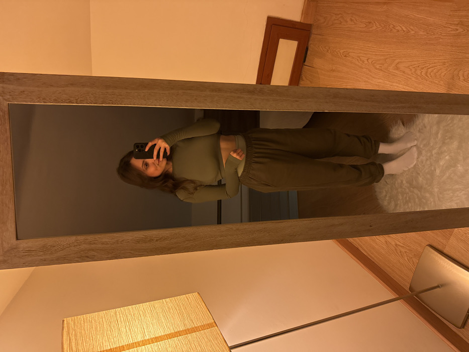 The image displays an indoor scene featuring a female individual captured in the reflection of a full-length mirror with a light wooden frame. The individual, with dark brown hair and a slight smile, is holding a dark-colored smartphone to her face, indicating the capture of a self-portrait. She is dressed in a long-sleeved, olive-green cropped top, revealing her midriff, with a lighter green waistband visible below. She wears dark olive-green sweatpants and white socks. She stands on a white, shaggy rug covering a portion of a light wooden floor. To the immediate left of the mirror, a tall floor lamp with a rectangular, textured beige shade and a brushed metallic base emits warm-toned artificial light. In the background, reflected on the right side of the mirror, a dark brown wooden frame leans against a light-colored wall. The specific location within Cava de' Tirreni, Italy, cannot be determined from the image content.