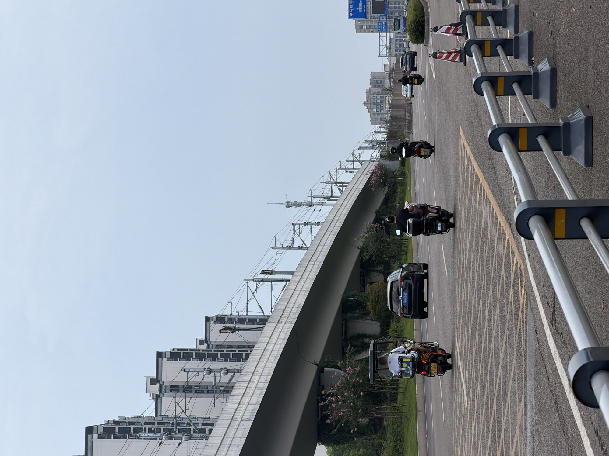 The image shows a street scene in Jiangjin District, Chongqing, China, with a highway overpass dominating the background. Several residential buildings are visible alongside the overpass. On the street, there are multiple vehicles, including a tricycle, several motorcycles, and cars. People are riding the vehicles. The sky is overcast. Traffic cones and guardrails are present on the side of the road.