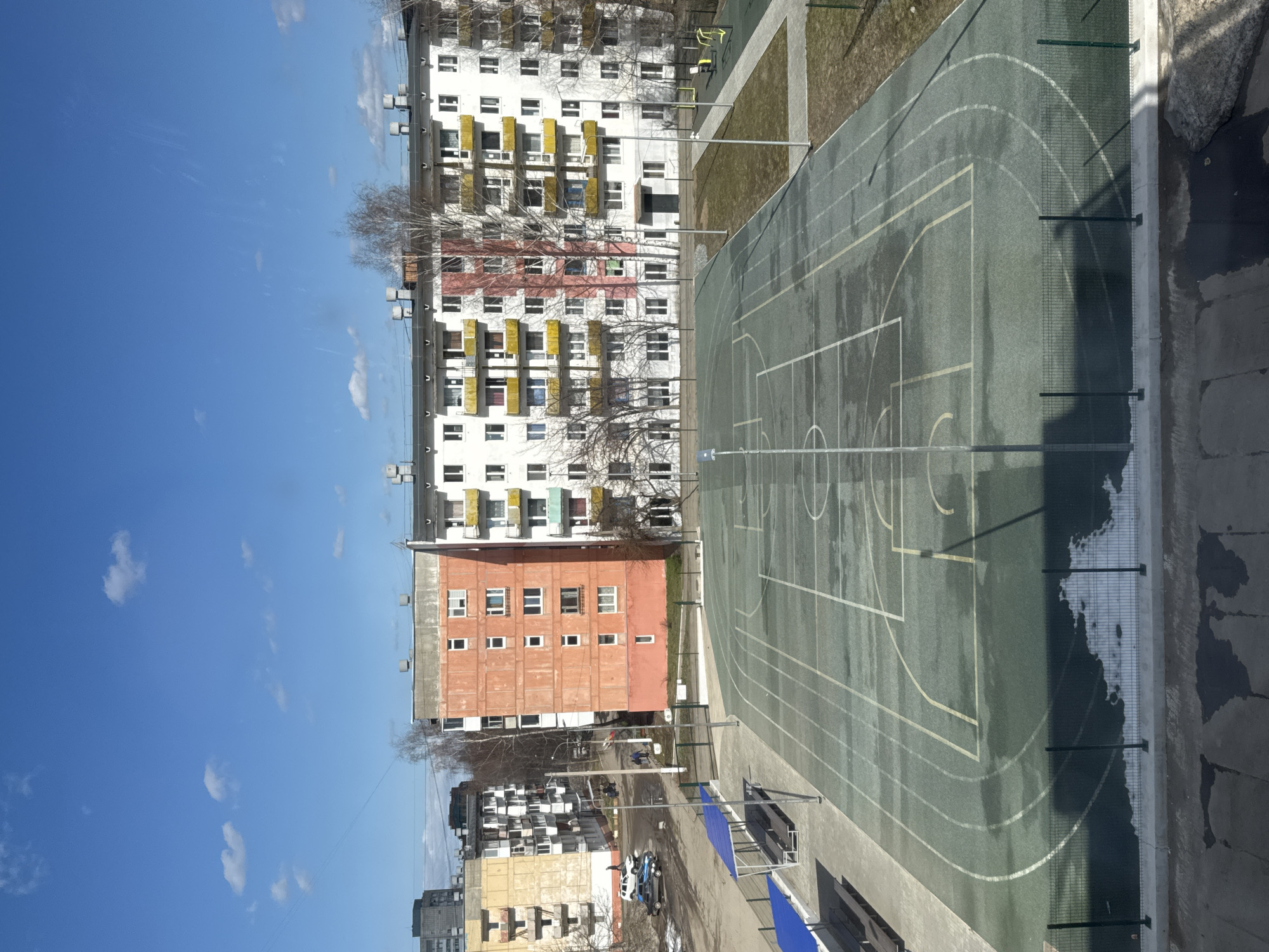 The image shows an elevated view of a sports ground in Chaikovsky, Russia, with a basketball court marked with lines and running tracks. The court is surrounded by a green fence. Adjacent to the court is a grassy area with some playground equipment. In the background, there are several multi-story residential buildings, varying in color from white with yellow balconies to orange. The sky is clear blue with scattered clouds. Below the sports ground, some parked cars are visible.