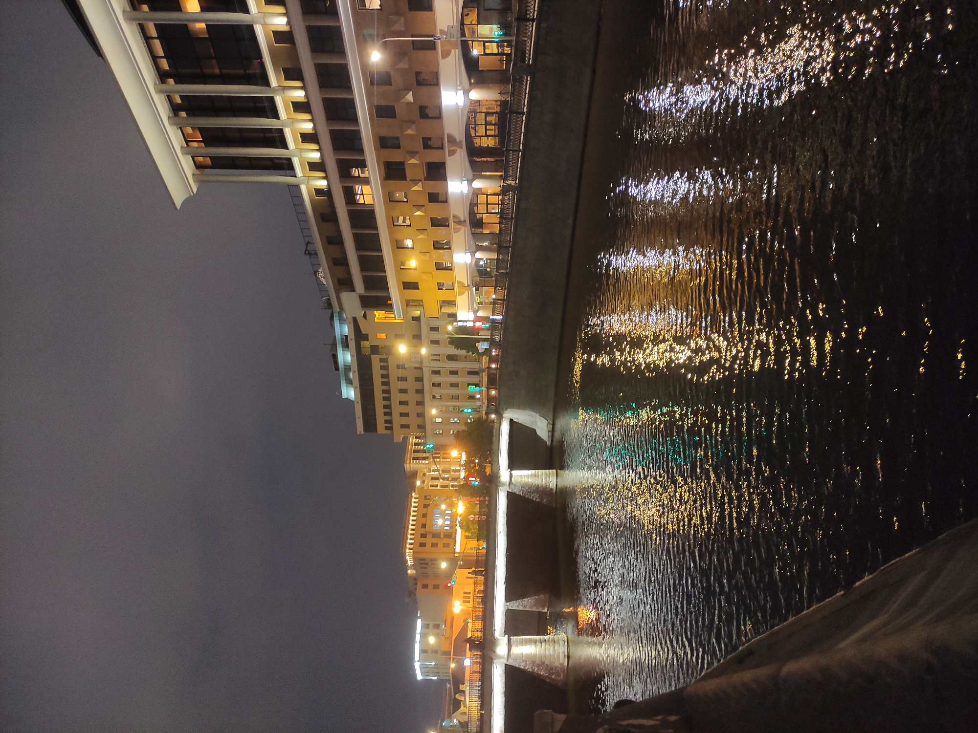 Here is a description of the image:
The image shows a nighttime view of the Moscow River in Russia. The river reflects the lights of the city, including buildings and street lamps. A bridge spans the river. Buildings are seen on the left of the image. The sky is dark and overcast. The image is taken from a low angle, near the river's edge.