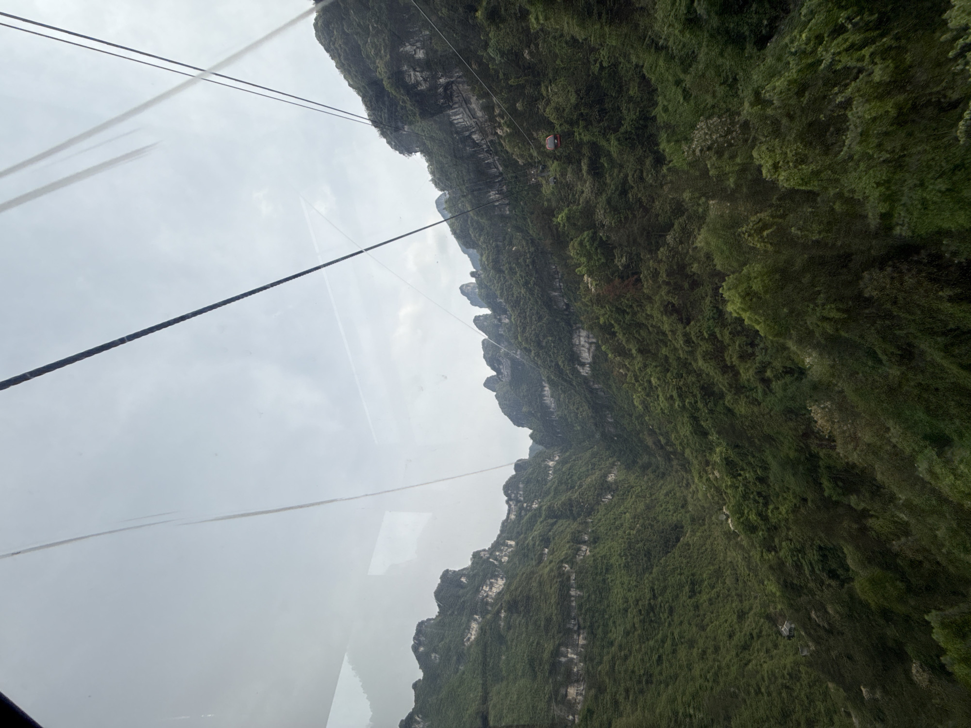 The image shows a view of a mountain range from a cable car in Zhangjiajie, China. The mountains are covered in dense green vegetation, and the sky is overcast. A cable car is visible in the distance. The foreground shows the cable and the interior of the cable car with streaks on the glass.