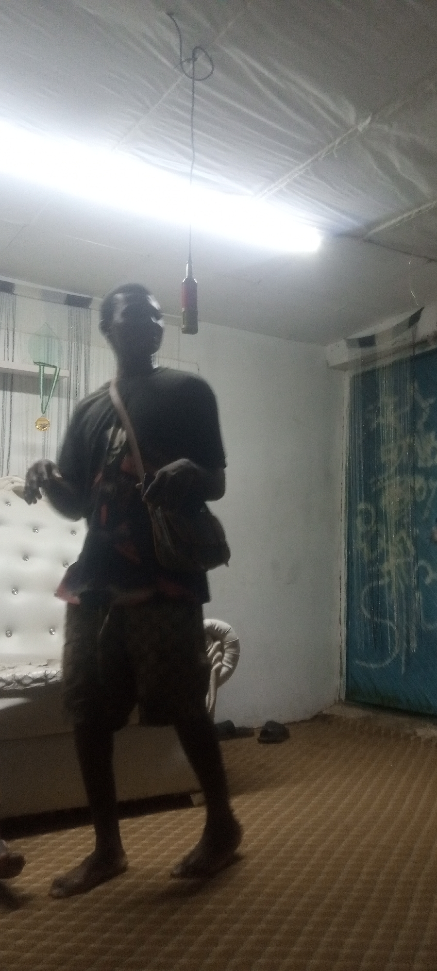 A dimly lit interior space contains a man, photographed at approximately eye-level. The man, wearing a dark-colored T-shirt and shorts, is standing barefoot on a patterned tan carpet. He has a bag slung over his shoulder. Behind the man, a white tufted piece of furniture, possibly a couch or headboard, is visible. A doorway is seen on the right side of the image; the door itself is painted blue and bears some graffiti. Overhead, a fluorescent light fixture dominates the center of the ceiling, with a hanging item suspended by a wire nearby. A medal is hung on a shelf above the white furniture. The city and state are unknown.