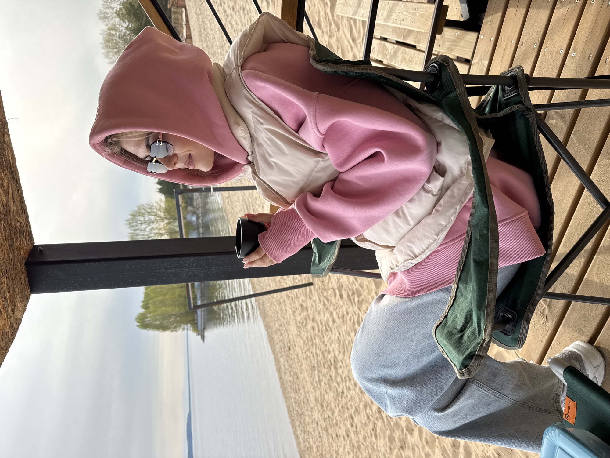 A person wearing a pink hooded sweatshirt, a cream-colored vest, and gray sweatpants sits in a green folding chair on a wooden platform. The person is also wearing sunglasses and holding a dark-colored cup. The platform overlooks a sandy beach and a body of water, possibly a lake or a sea, under a cloudy sky. Trees are visible in the background across the water. A dark beam supports the platform structure on the left side of the frame.