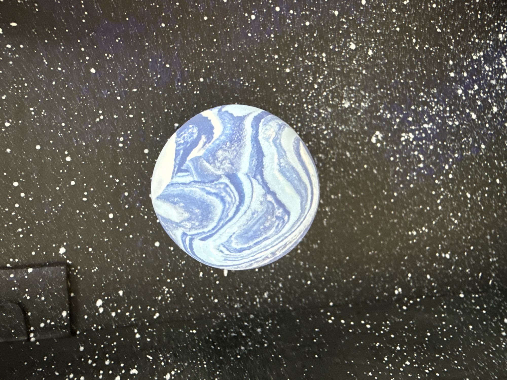 A circular object with swirling patterns of light blue and white resembling a planet floats in a black expanse speckled with white dots resembling stars. A dark, rectangular shape is visible in the lower left corner. No identifiable location or people are present in the image. The scene evokes a cosmic or space-themed setting.