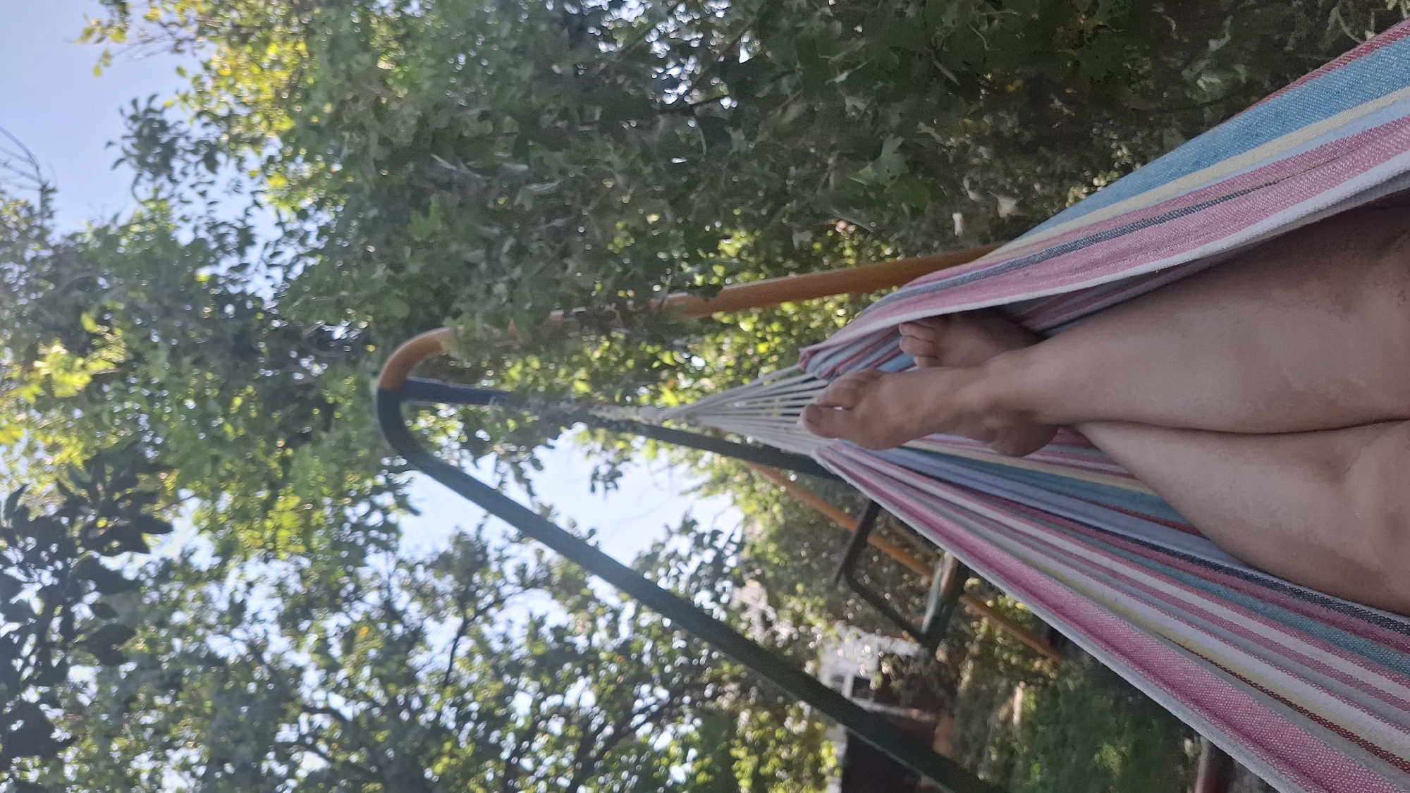 The image shows a person's legs and feet lying in a hammock. The hammock is made of colorful striped fabric and is suspended between two poles, which appear to be part of a swing set. The background consists of lush green trees and a clear blue sky, suggesting an outdoor setting, possibly a backyard or park. The sunlight filtering through the leaves indicates it is daytime. The person's legs are crossed, and their bare feet are visible at the top of the frame. The overall impression is one of relaxation and leisure in a natural environment. The location, Шұбарсу, Kazakhstan, implies this scene is set within a residential area or a public space in that region.