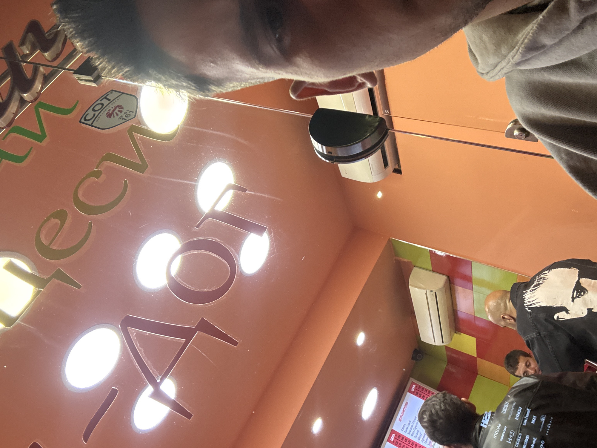 The image shows a person taking a photo from a high angle inside what appears to be a fast-food restaurant in Sofia, Bulgaria. The ceiling is orange with cut-out letters illuminated from behind, spelling out what seems to be the name of the place. A logo with "COT" is also visible on the ceiling. The interior has an air conditioning unit and other light fixtures mounted on the ceiling. Below, there are customers waiting in line, wearing jackets. A menu board is partially visible. The background features a tiled wall with red, yellow and green colors.