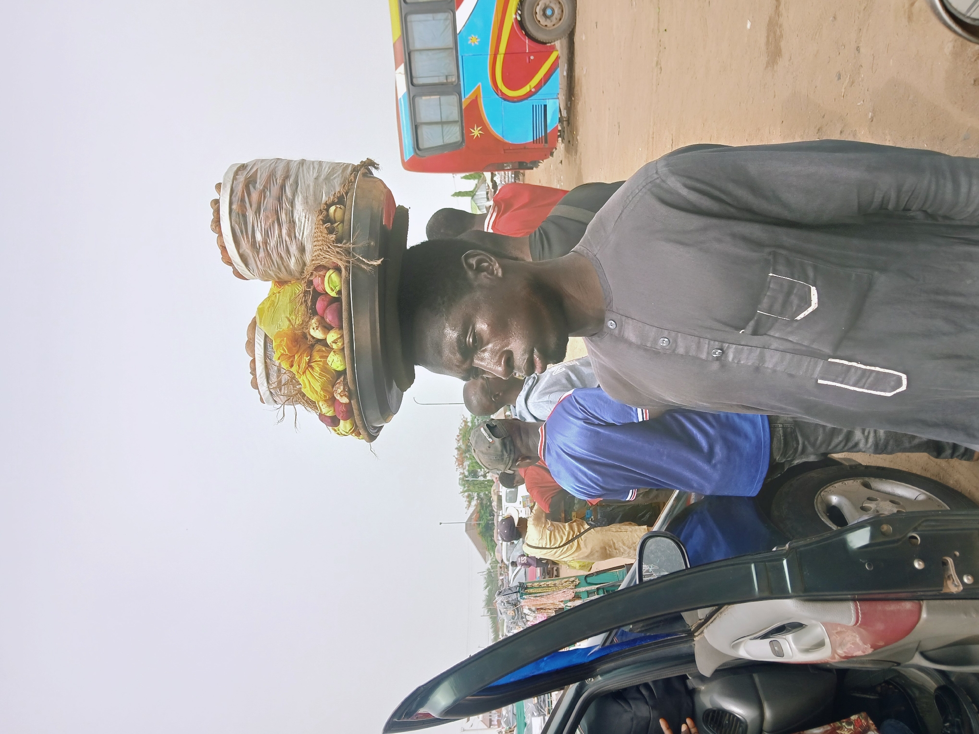 A man is standing near a car with an open door, balancing a tray of fruit and nuts on his head. He is wearing a dark shirt, and sweat is visible on his face. Other people are nearby, some wearing brightly colored shirts. A bus with colorful paint is in the background. The location appears to be a busy street or marketplace.