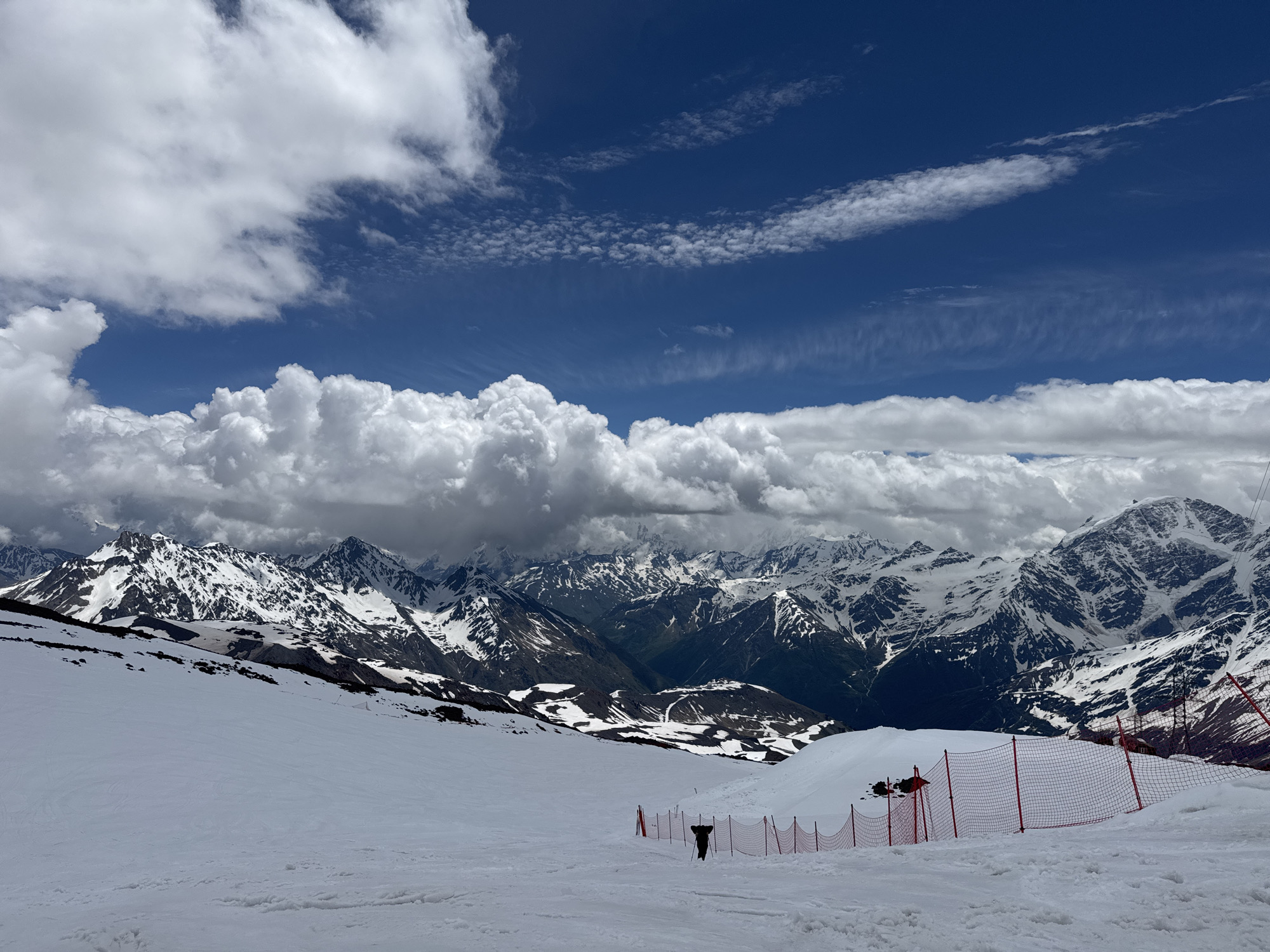 An extreme wide shot captures a vast, snow-covered mountain landscape under a dynamic sky filled with white, fluffy clouds against a deep blue backdrop. In the foreground, a gentle slope of pristine white snow stretches out, marked by faint ski tracks and a sparse scattering of dark rocks. A bright orange safety net with white poles runs along the middle ground, delineating a boundary. A solitary figure, dressed in dark clothing and appearing to hold ski poles, stands near the net, facing away from the camera. Beyond the net, the landscape transforms into a dramatic panorama of jagged, snow-capped mountain peaks receding into the distance. The peaks are a mix of white snow and dark, rocky outcrops, with shadows adding depth and contrast. In the far background, the mountains become softer and more hazy, hinting at further ranges. The sky above is a vibrant blue, interrupted by large, billowing white clouds that cast shadows on the mountains below, suggesting a transition in weather. The image is taken from a high vantage point, likely a ski slope, overlooking the majestic mountainous terrain of Terskol, Russia.