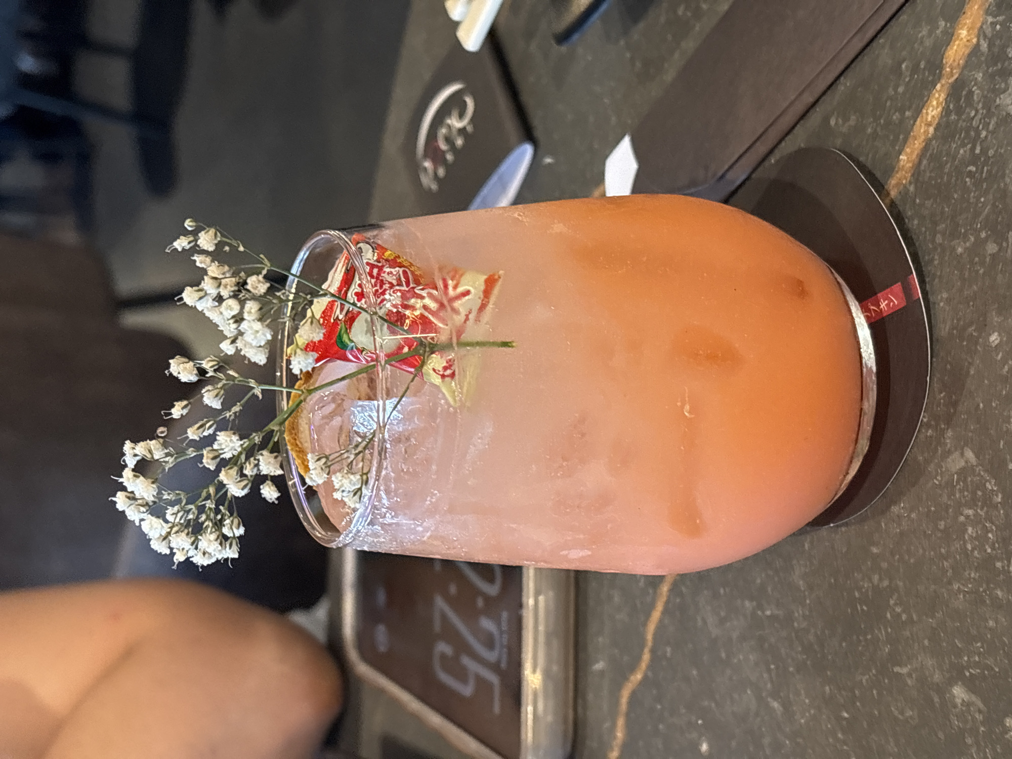 A horizontally-oriented medium shot features a cocktail in a clear glass, adorned with white baby's breath flowers and an orange garnish, set on a coaster on a table. The cocktail has a pinkish-orange hue. The coaster is black with a thin silver rim. To the left of the cocktail, a person's shoulder is visible. Behind the cocktail, there is a phone with "2:25" displayed on the screen, and parts of a black tabletop setting. The surface of the table has a dark grey, textured appearance with a vein of gold running across it. The location is unclear.