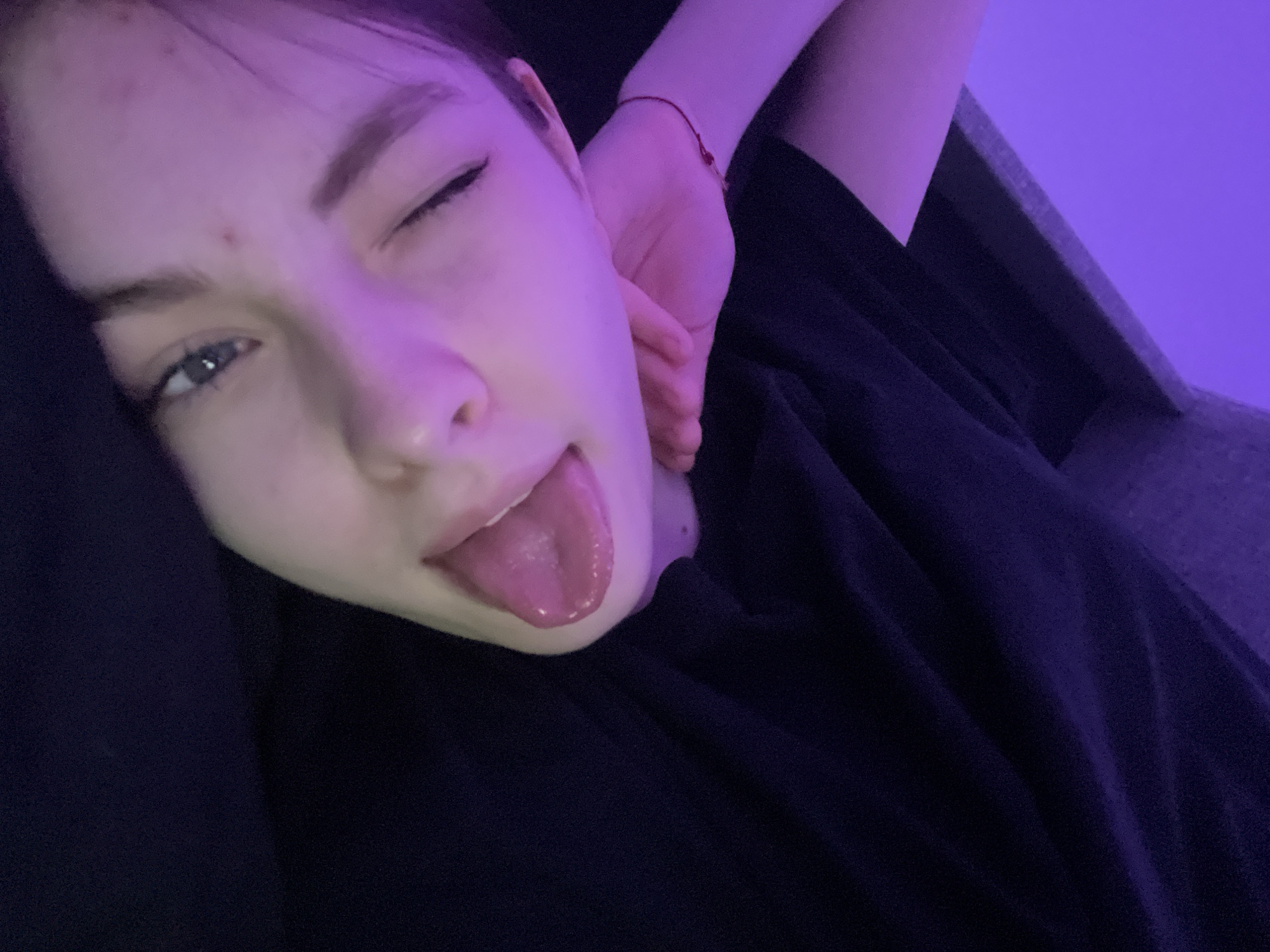 A person with fair skin and blue eyes is winking and sticking their tongue out. The person is wearing a black top and has a red bracelet on their wrist. The background is a purple hue, possibly from lighting, and includes a dark gray sofa or chair. The person appears to be lying down or reclining. The location is not identifiable as Saint Petersburg, Russia, based on the provided image.