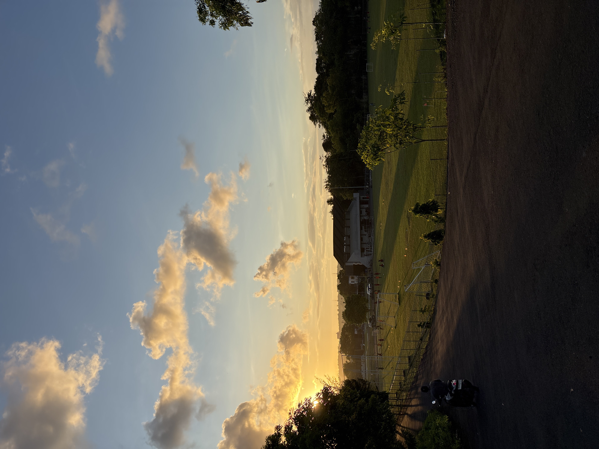 The image depicts a scene at dusk in Kuta Selatan, Indonesia. The sky is visible with hues of blue, yellow, and orange, punctuated by clouds. A sports field is adjacent to a road, bordered by a fence. A person is near a vehicle parked along the road. The environment includes trees, grass, and a building further into the scene. There are objects present such as soccer goals on the field.