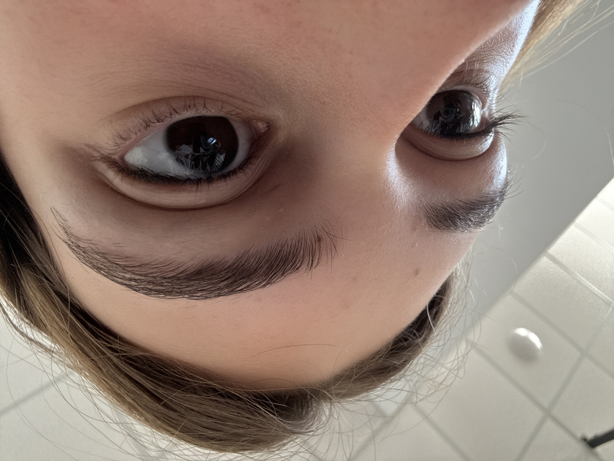 A close-up, angled view of a person's face focusing on their eyes and eyebrows. The person has brown eyes and dark brown eyebrows. The skin tone is fair. The perspective is looking up at the person, distorting the facial features. The background is blurry and includes a portion of light brown hair, a white surface with a light reflecting off it. The location is not discernible from the image.