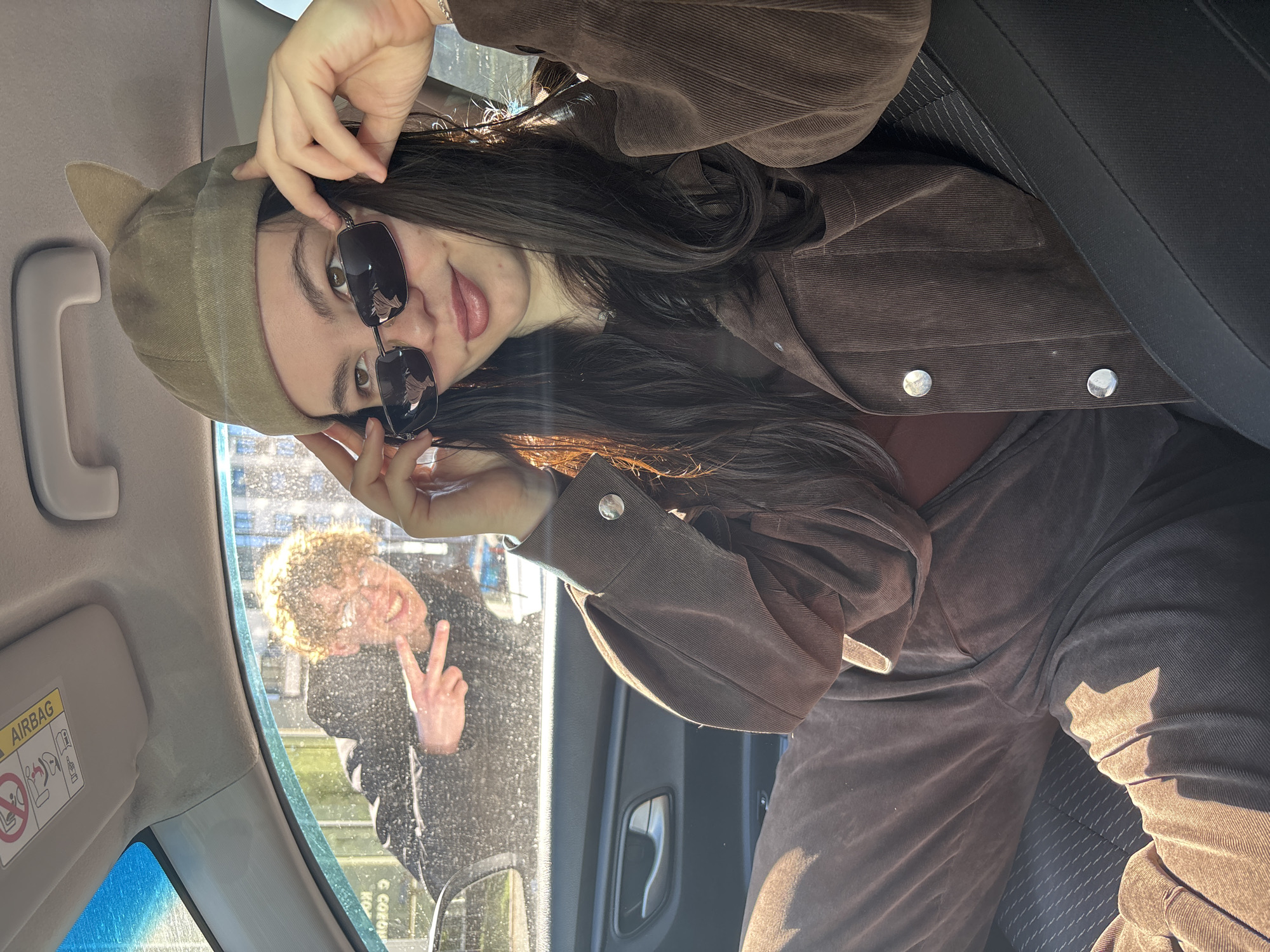 A young woman with dark hair, wearing a brown corduroy jacket and matching pants, sits in the driver's seat of a car. She is wearing sunglasses and a beige cat-eared hat. Through the car window, a young man with curly hair is visible, holding up a peace sign. An "AIRBAG" warning sticker is visible on the car's sun visor. The picture was taken in the city of Perm, Russia.