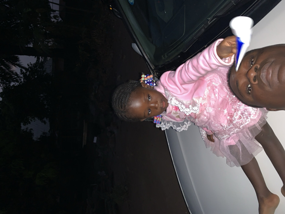 A young girl in a pink dress with colorful hair ties stands on a white car hood alongside an adult male. She holds an object resembling a tube of cream in her hand. Her pink dress is frilly and adorned with lace. The background shows what looks like trees and a building, suggesting an outdoor setting, possibly at dusk or night due to the darkness surrounding the scene.