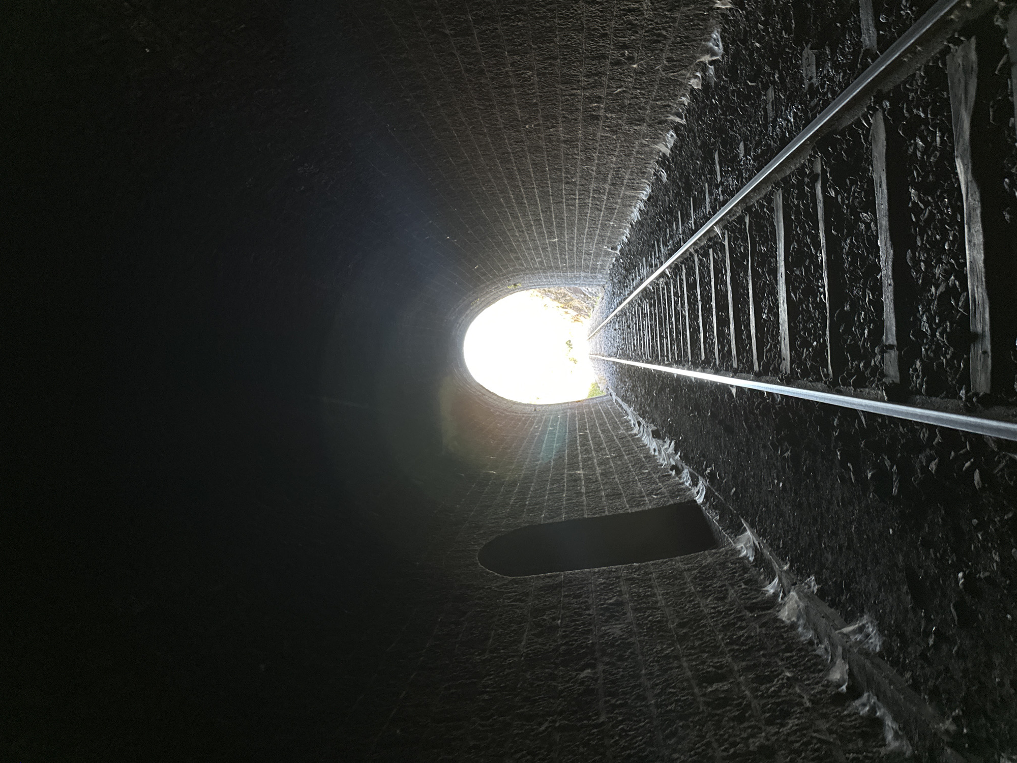 The image depicts the interior of a train tunnel with a railway track leading towards a brightly lit opening at the end. The tunnel walls are constructed of rough-hewn, dark stone blocks. On the left side of the tunnel, there is a semicircular arched opening, recessed into the wall. The ground of the tunnel is covered with ballast and debris. The railway track consists of two metal rails supported by wooden sleepers. The opening at the end of the tunnel reveals bright daylight, with hints of greenery visible outside. The scene is captured from a low-angle perspective, looking down the track and towards the light.
