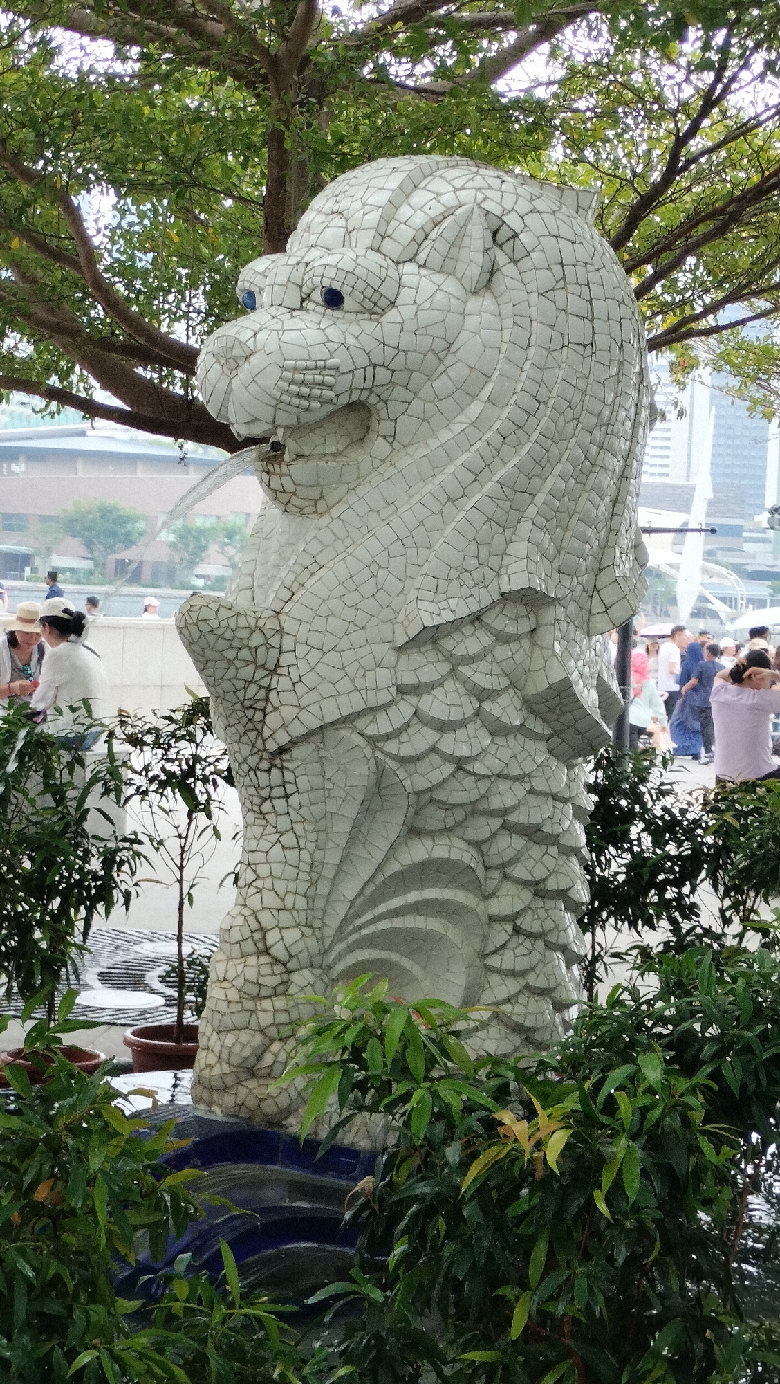 The image shows the Merlion statue, a prominent landmark in Singapore, located at Merlion Park along the Marina Bay waterfront. The statue is a mythical creature with a lion's head and a fish's body, and it is spewing water from its mouth, creating a fountain effect. The statue is covered in white mosaic tiles, with blue mosaic tiles for its eyes.
In the foreground, lush green bushes and small trees partially obscure the base of the statue and the water basin. Behind the statue, a checkered pattern is visible on the ground, possibly a pathway or plaza. In the mid-ground, several people are present. On the left, two women, one wearing a straw hat, are seated on a white ledge, appearing to be in conversation. Further to the right, a crowd of people are milling about, some standing and others walking. They are dressed in casual attire.
In the background, a tree with dense green foliage branches out over the statue. Beyond the people and the park, modern city buildings, including skyscrapers, are visible, indicating an urban environment. The sky appears overcast. The overall scene depicts a public space in Singapore, likely a tourist attraction, with the iconic Merlion statue as the central focus.