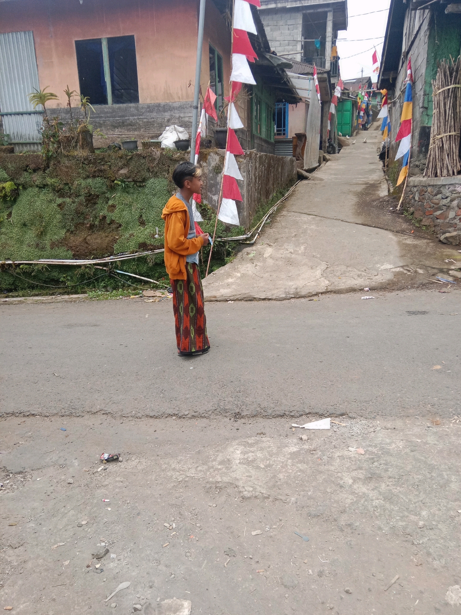 This image captures a street scene in Batur, Indonesia, possibly during a cultural celebration given the numerous red and white flags lining the pathway. The foreground is dominated by an asphalt road, showing signs of wear and some scattered debris. A young boy, likely of Indonesian descent, stands on the right side of the road, facing away from the camera and looking down the street. He is wearing an orange hooded jacket over a gray t-shirt and a long, patterned sarong-like garment that extends to his feet. His hair is short with a blondish-brown streak at the front. The street itself slopes gently upwards into the distance, flanked by residential buildings constructed from a mix of concrete, brick, and corrugated metal. Greenery and some potted plants are visible on a raised embankment to the left of the boy. In the background, the street opens up into a wider area with more houses and flags, suggesting a more populated part of the settlement. The overall impression is of a modest, residential street in a tropical or subtropical climate.