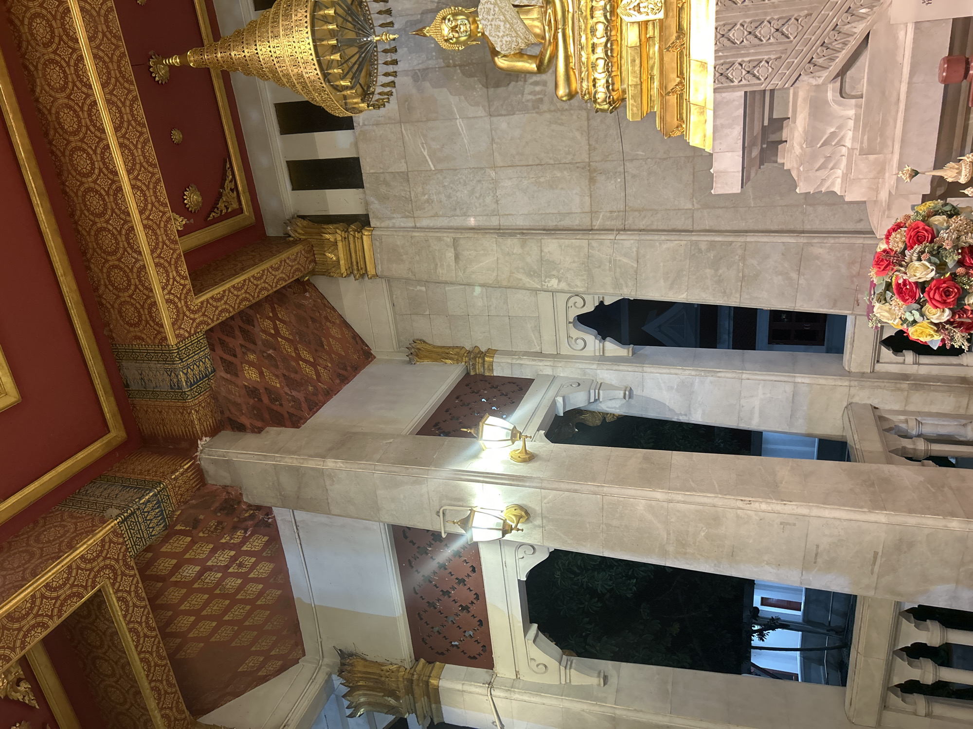 This is a photograph of a Buddhist temple interior in Tha Khlong Town Municipality, Thailand. The scene is dominated by a large, ornate golden statue of Buddha seated on a tiered pedestal. The statue is adorned with a white and gold sash. Above the Buddha, a golden tiered umbrella-like structure hangs from the ceiling.
The ceiling itself is a striking feature, painted in a deep red with intricate gold patterns and decorative borders. Large, polished stone columns, likely marble, support the ceiling and frame the space. Two wall-mounted lanterns with white glass shades and gold fixtures are visible on one of the columns, illuminating the area.
In the foreground, to the lower right, a bouquet of colorful artificial flowers, including red, yellow, and white roses, is placed near the base of the Buddha statue. A small tiered, spire-like ornament is also visible. Through an arched opening to the left, glimpses of trees and parts of a building with red doors and white trim can be seen in the exterior darkness. The walls of the temple appear to be constructed from large, light-colored stone tiles. The overall atmosphere is one of reverence and grandeur, characterized by the rich colors, intricate details, and the presence of the Buddha statue.
