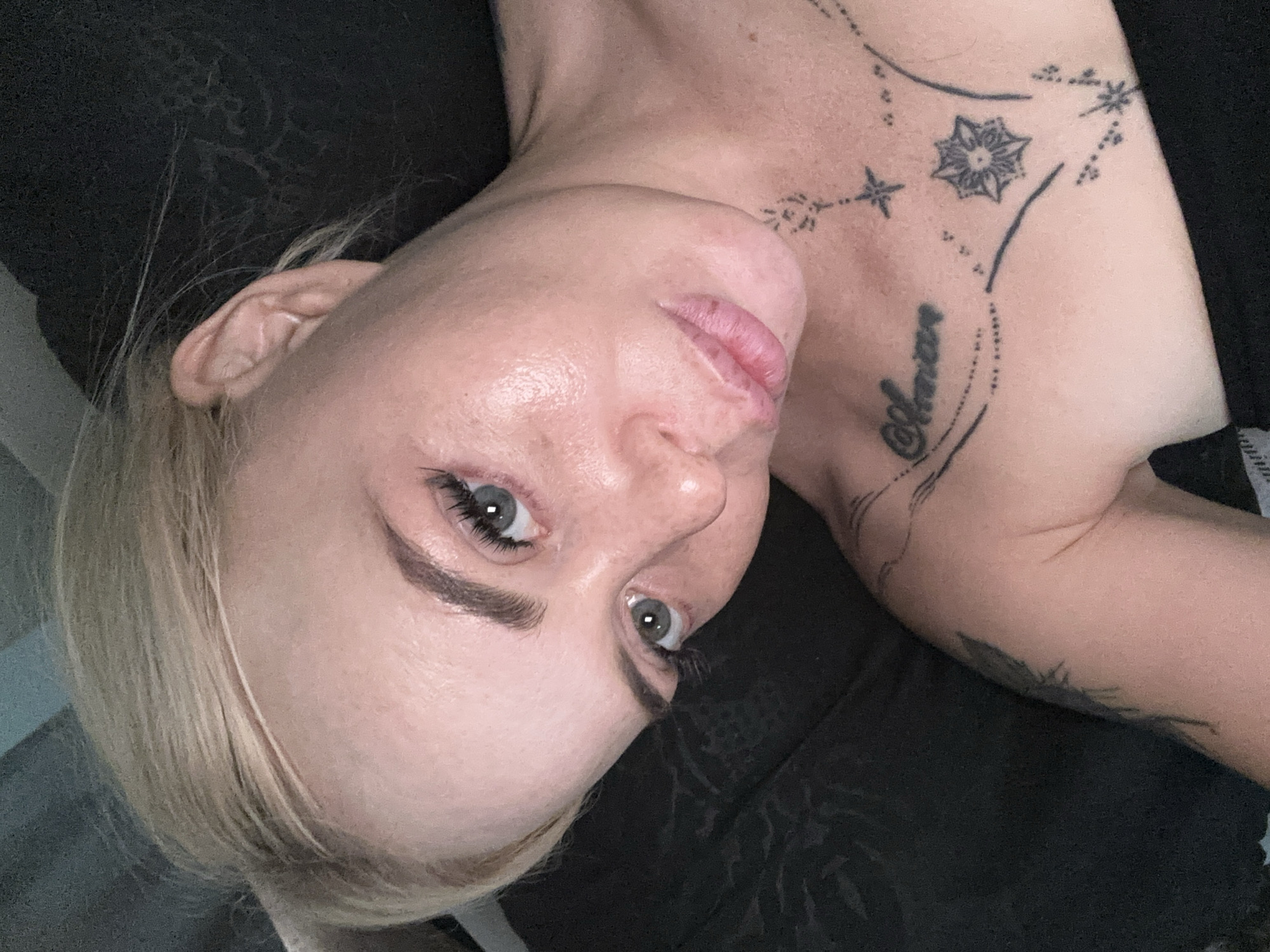 A close-up, overhead shot shows the upper body of a fair-skinned woman with blonde hair. She is lying down, with her head tilted to the left. Her eyes are open and looking slightly upward. She is wearing makeup, including mascara and possibly eyeshadow, and her eyebrows are well-defined. She has noticeable eyelashes. Her lips are slightly parted. The woman has tattoos on her chest and shoulder. One tattoo on her chest appears to be script with an asterisk and starburst designs. Another tattoo on her arm, partially visible, seems to be of a winged creature. The background is dark and appears to be a textured fabric, possibly a blanket or clothing. The lighting is soft and highlights the sheen on her skin.