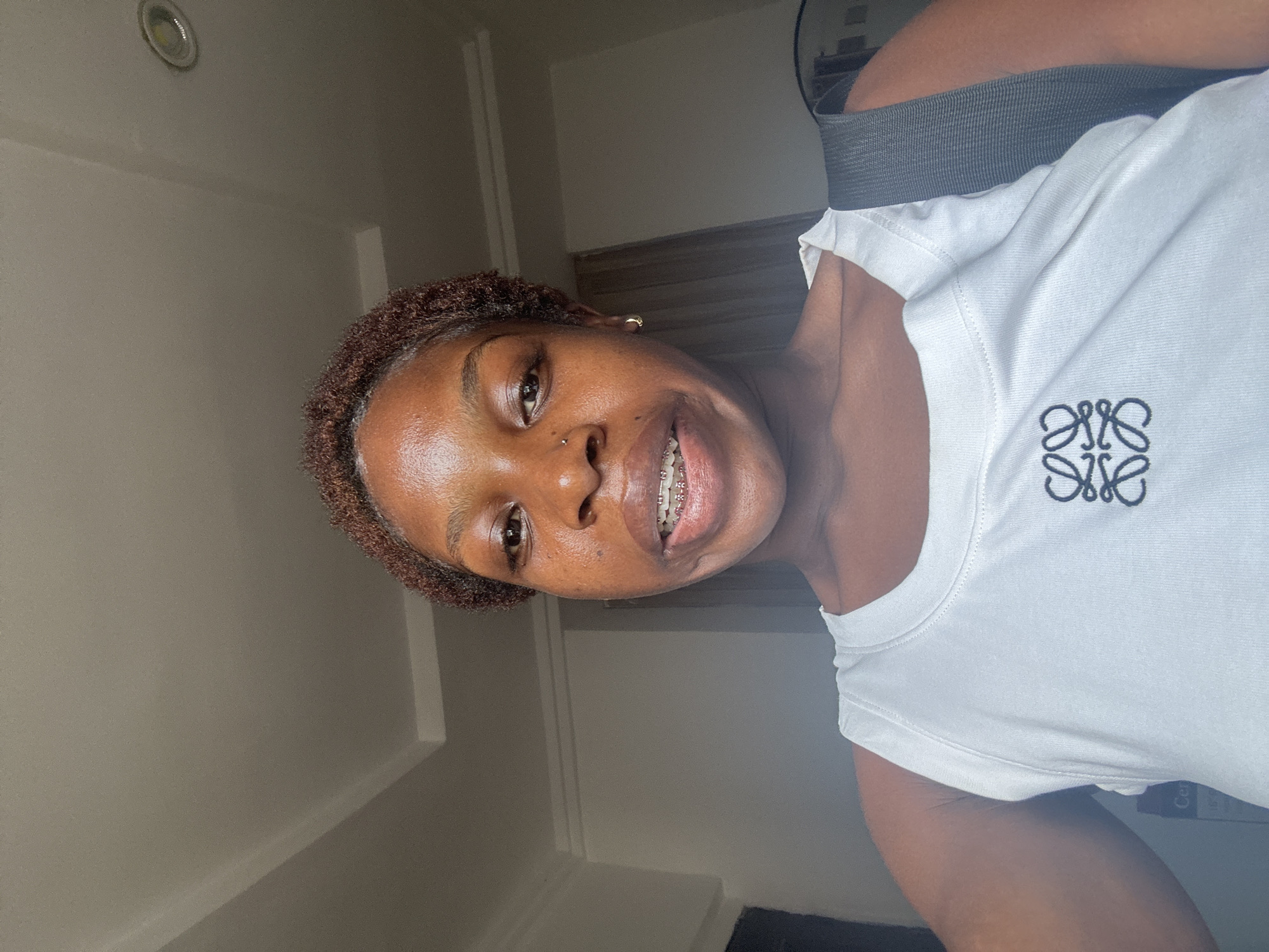 A young dark-skinned woman with short, curly brown hair is featured in a close-up, tilted selfie. She is smiling broadly, revealing dental braces, and looking directly at the camera. Her facial features include a small nose stud and a subtle gold hoop earring in her left ear. She is wearing a plain white t-shirt that features a distinctive black embroidered logo on the chest, identified as the Loewe Anagram (four interlocking L's).
The setting appears to be an indoor room with light-colored, possibly off-white or light grey, walls and a white ceiling. A recessed circular light fixture is visible on the ceiling in the upper left of the frame. Part of a dark, possibly grey, textured object or piece of furniture is seen behind her right shoulder. The lighting is bright and even, suggesting either natural daylight or well-lit artificial indoor lighting. The woman's expression conveys happiness and comfort. The image is oriented at an angle, characteristic of a spontaneous self-portrait. Based on the provided context, the photo was taken in Abuja, Nigeria.