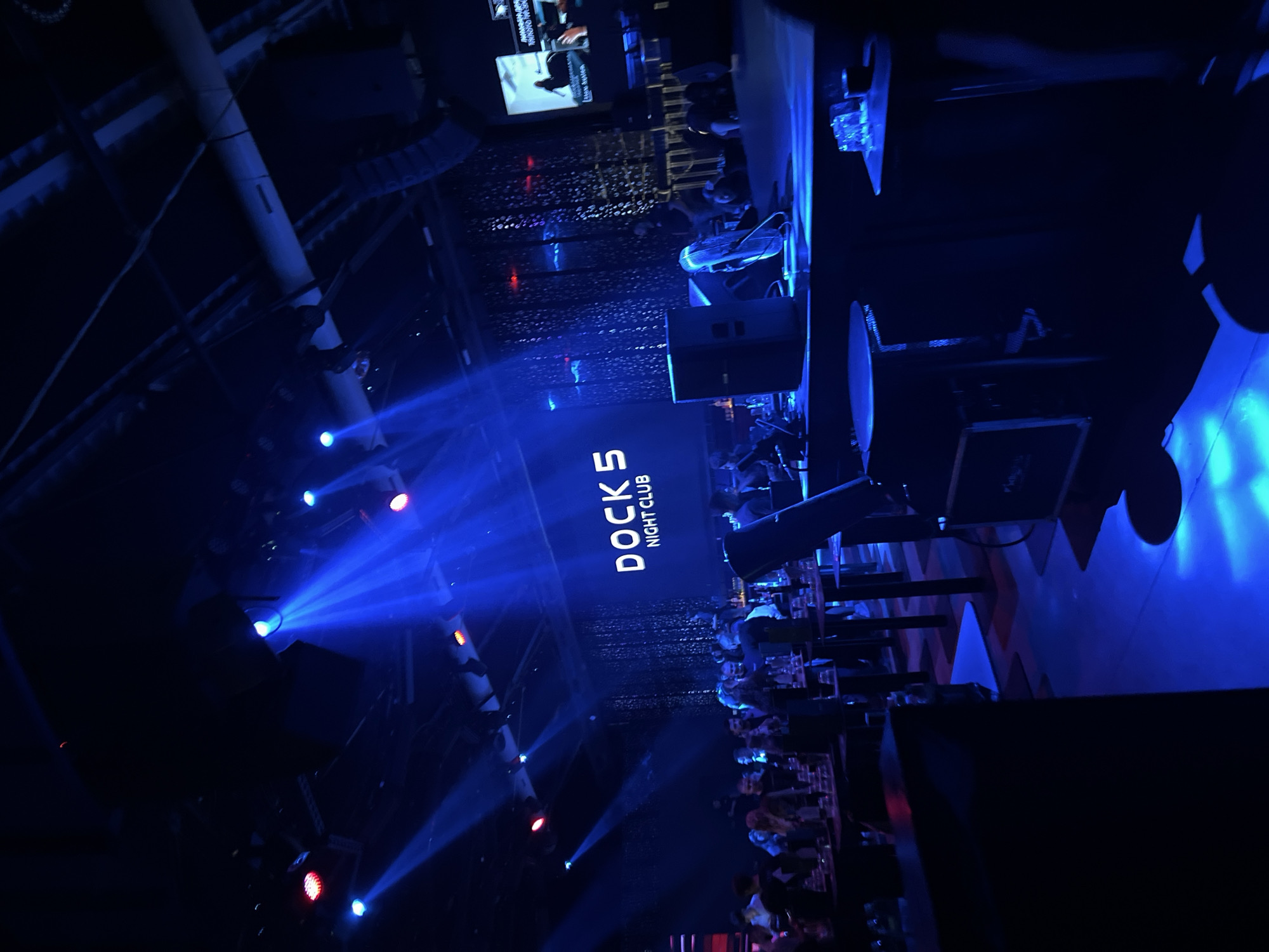 The image shows the interior of Docks 5 Nightclub, likely located in Burgas, Bulgaria. The club appears dimly lit with blue lighting and spotlights. People are present throughout the scene, mostly in seated areas. The club has a stage area. Various objects are visible, including tables, chairs, and audio equipment. The environment suggests an active nightlife venue with a focus on music and entertainment.