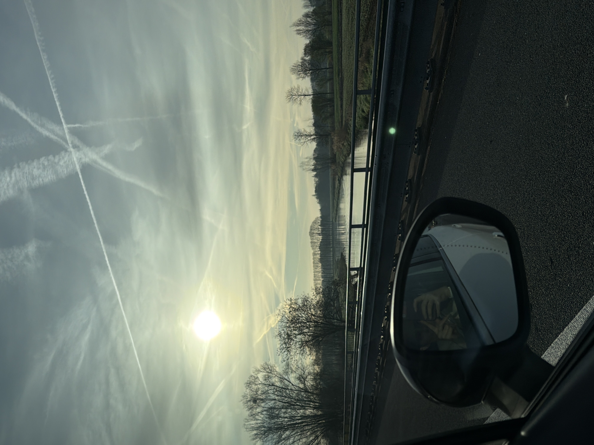 A view from a moving vehicle, likely on a bridge over a body of water. The location is near Heeswijk-Dinther, Netherlands, based on the environment, vegetation, and architectural elements along the river. The sky is overcast with numerous contrails crossing it, and the sun is visible. The side mirror of the car reflects the driver taking a photo, the asphalt of the road, the vehicle's exterior. The photograph is taken through the glass of the car window.